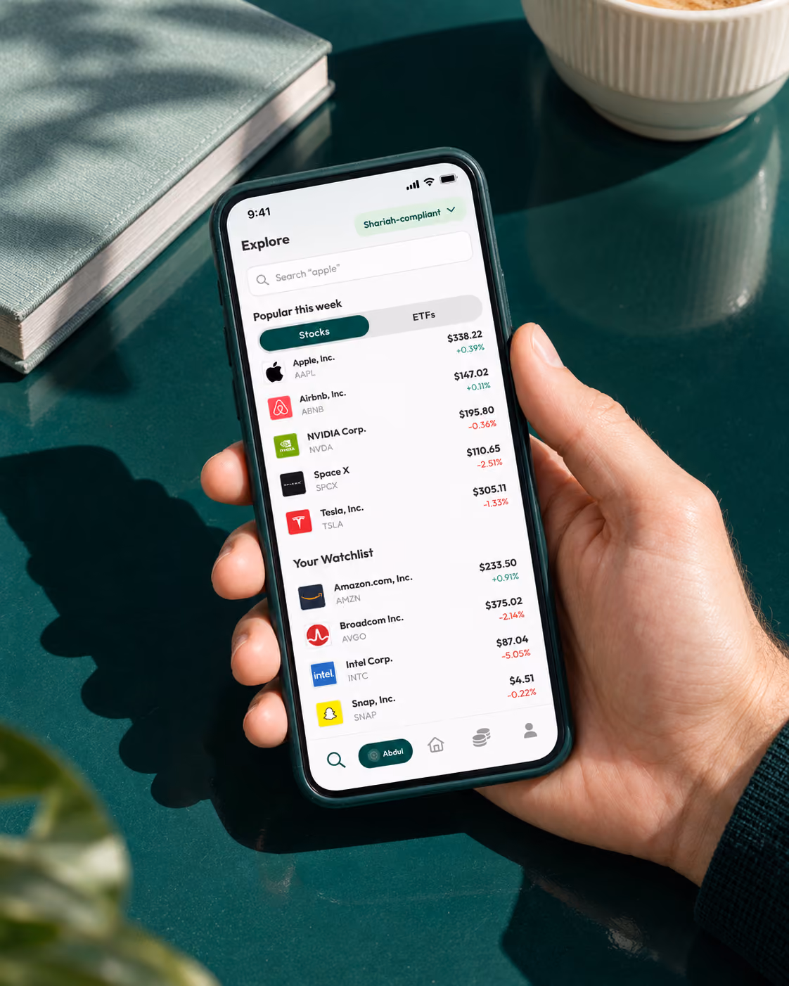 Hand holding a smartphone displaying a stock market app listing popular stocks this week including Apple, Airbnb, NVIDIA, SpaceX, and Tesla with their prices and percentage change, along with a watchlist featuring Amazon, Broadcom, Intel, and Snap.