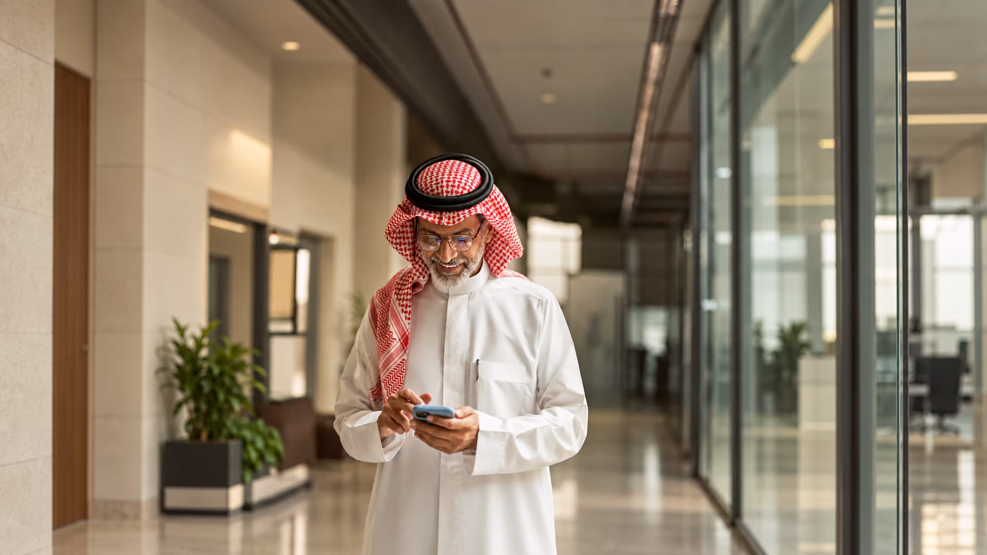 Smiling man wearing traditional Middle Eastern attire and red and white checkered headscarf, standing in a modern office corridor using a smartphone.