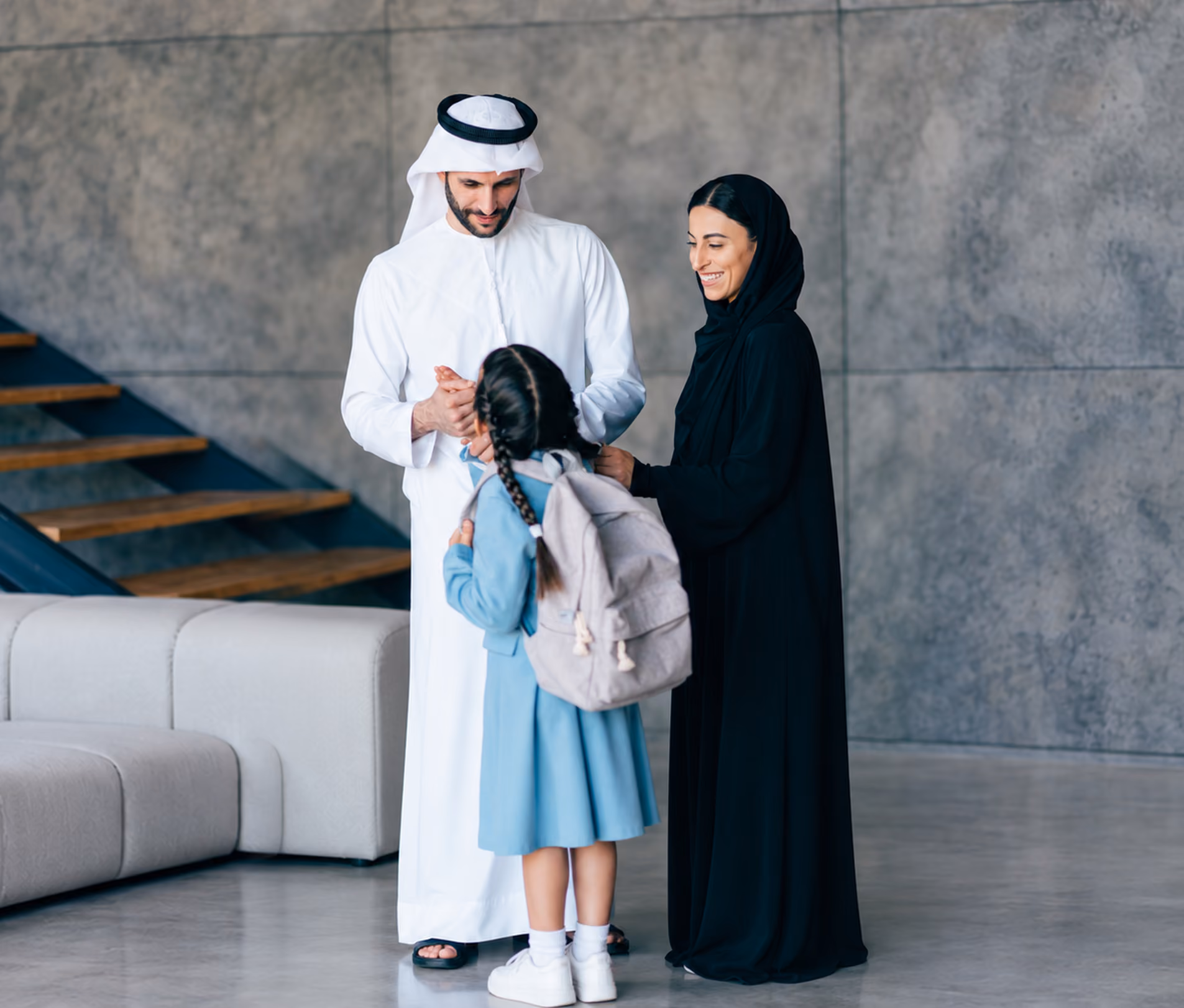 A man and woman in traditional Middle Eastern attire smiling at a young girl in a blue dress with a backpack inside a modern home with a gray concrete wall and a wooden staircase.