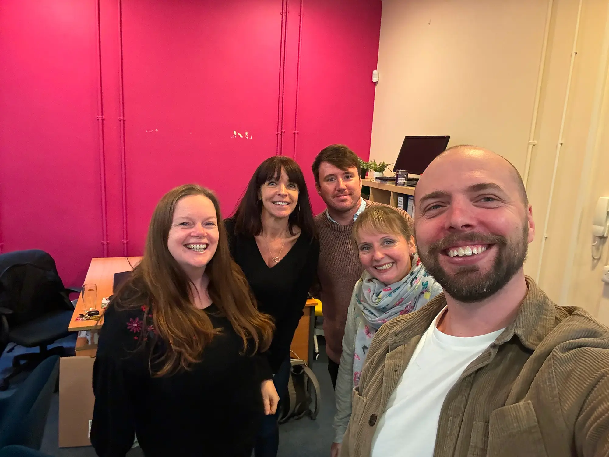 Group selfie of five smiling adults in an office with pink and beige walls.