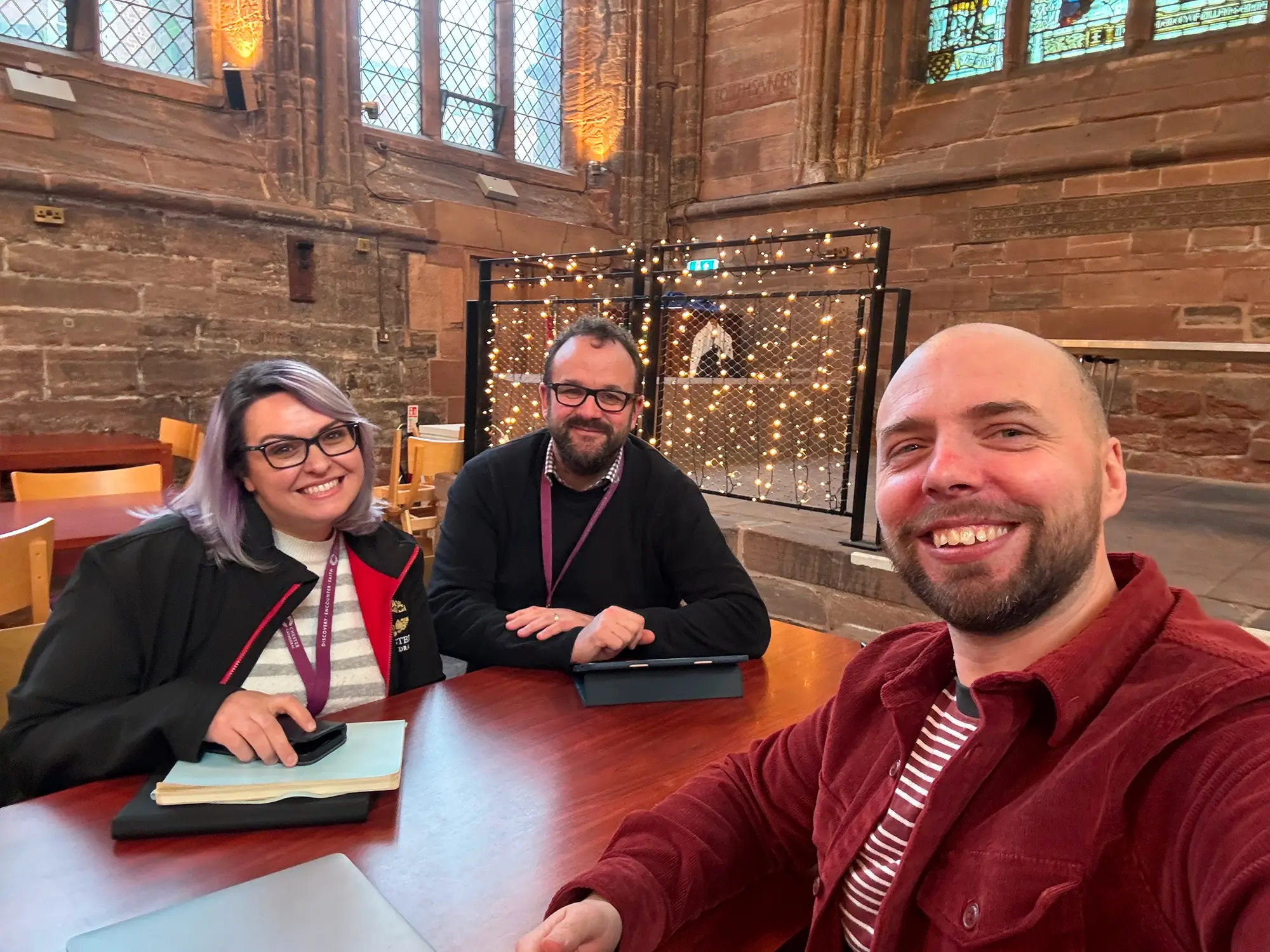 Three people smiling and sitting at a wooden table inside a building with stained glass windows and stone walls, with fairy lights in the background.