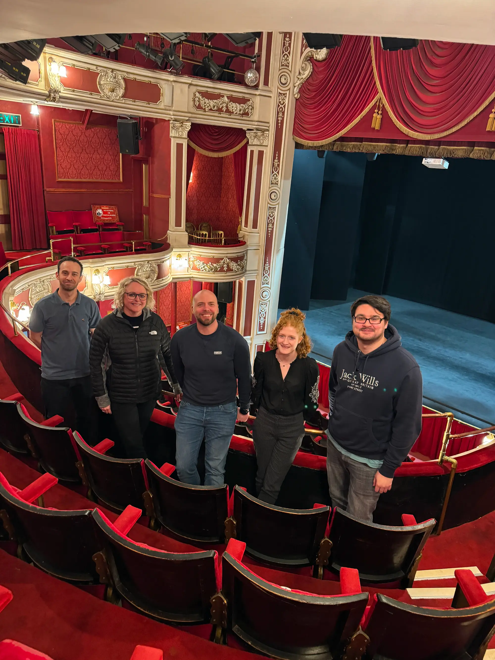 Five adults standing together smiling inside an ornate red and gold theater balcony with empty seats and a stage in the background.