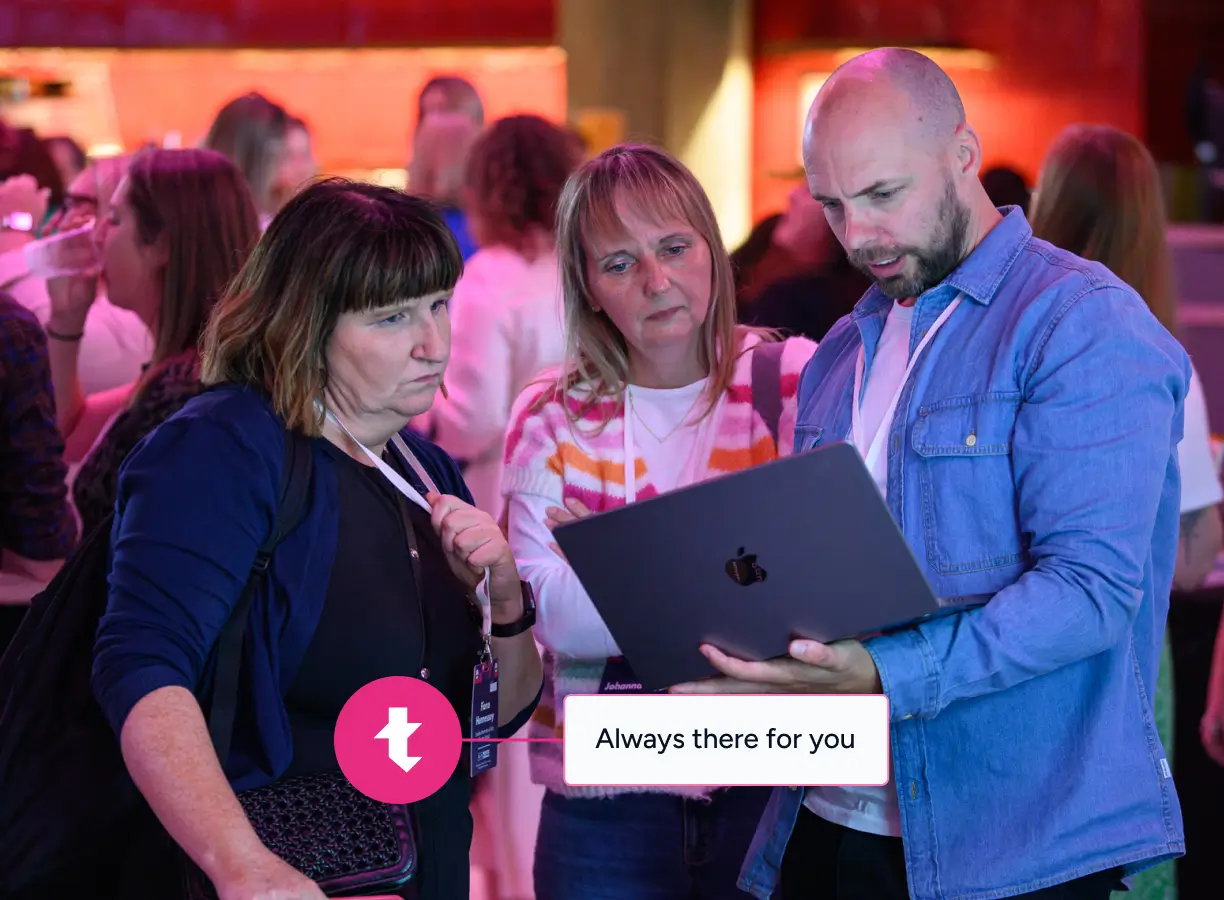 Three adults attentively looking at a laptop in a busy indoor setting with warm lighting.
