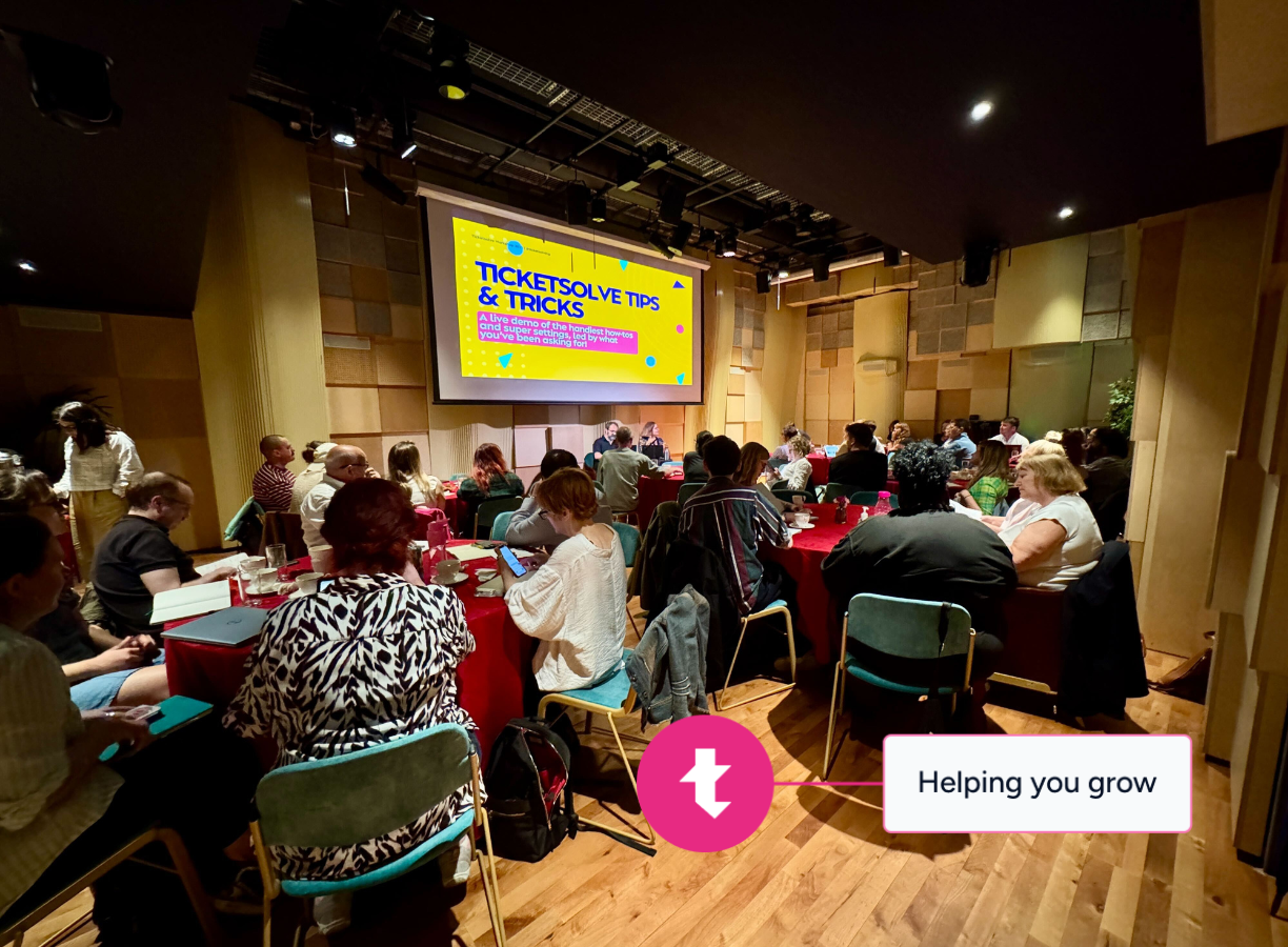 Group of people seated around tables in a conference room watching a presentation titled 'Ticketsolve Tips & Tricks'.