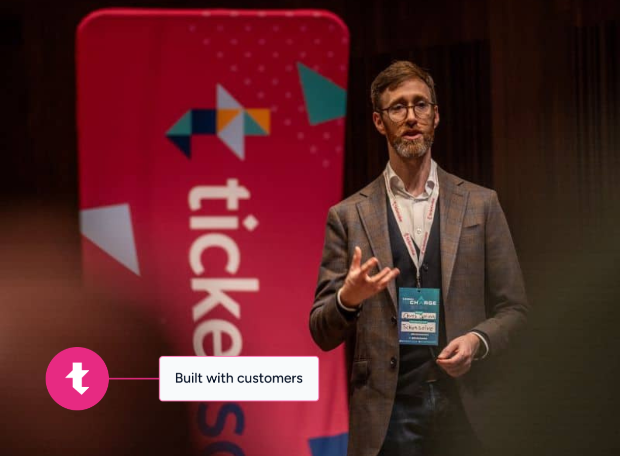 Man with glasses and a beard speaking on stage next to a red banner with the TicketSolve logo.