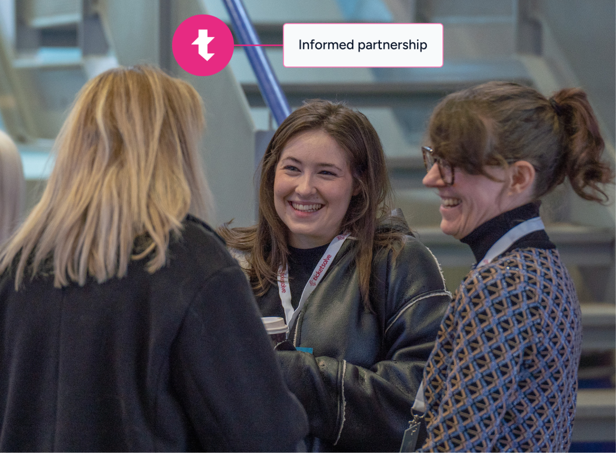 A man and woman wearing conference badges smile and talk while holding mugs, standing in front of a table with pink books and a pink 'ticket' backdrop.