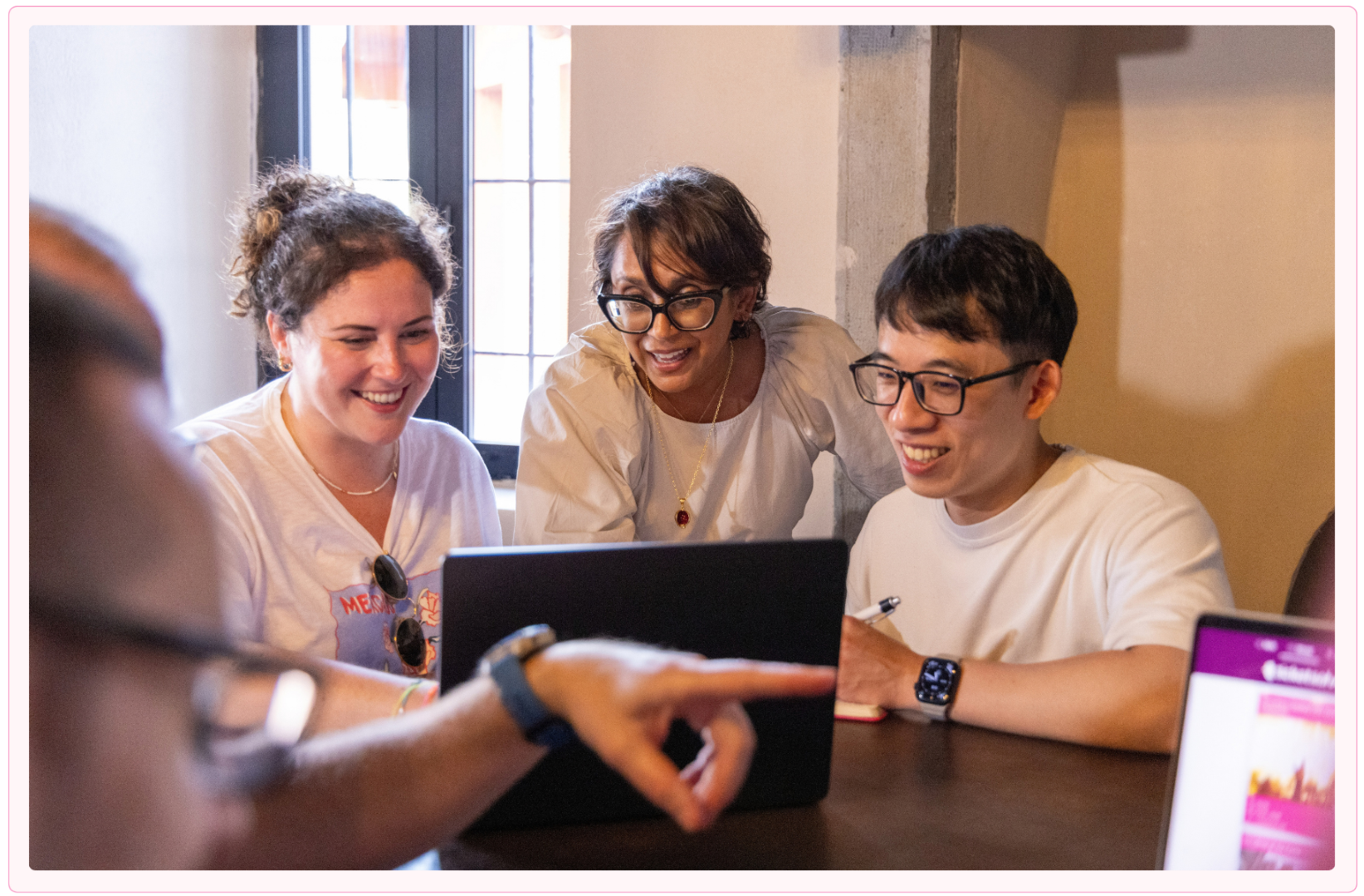 Three people smiling and working together on a laptop at a table with a window in the background, while a person in the foreground points at the screen.
