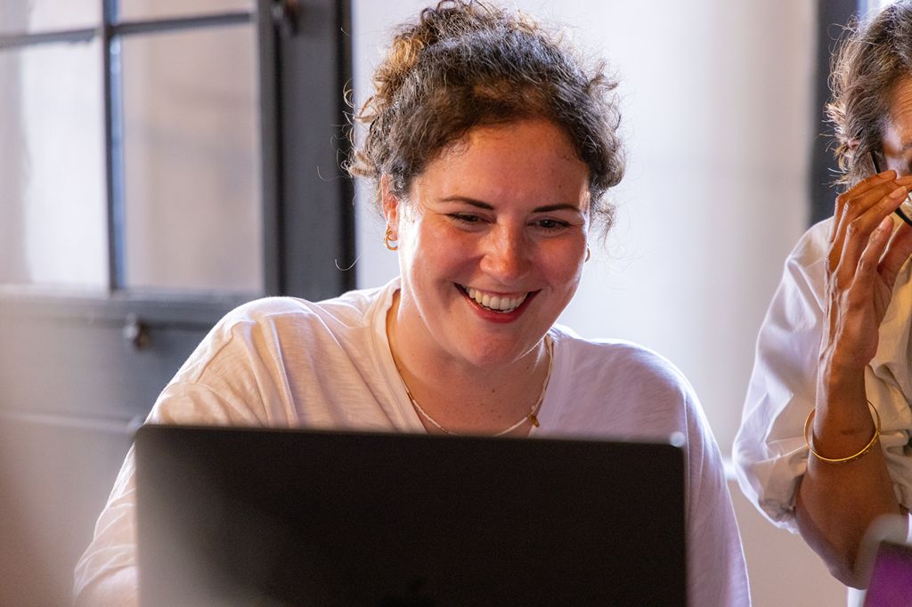 Smiling woman with curly hair working on a laptop in a softly lit room, next to another person partially visible on the right.