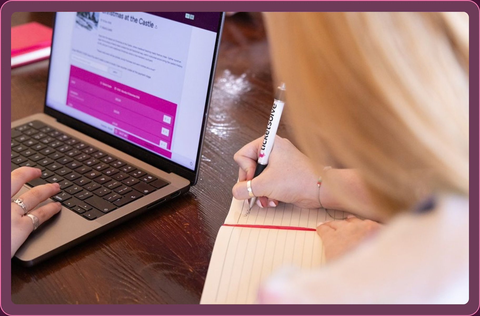 Person working at a laptop with a webpage open, writing notes in a lined notebook using a pen labeled 'TicketSolve'.