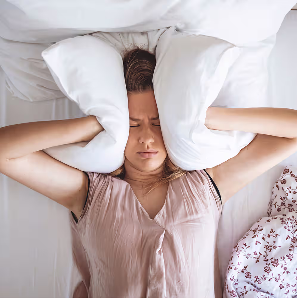 Mulher deitada na cama enquanto pressiona uma almofada em redor da cabeça.