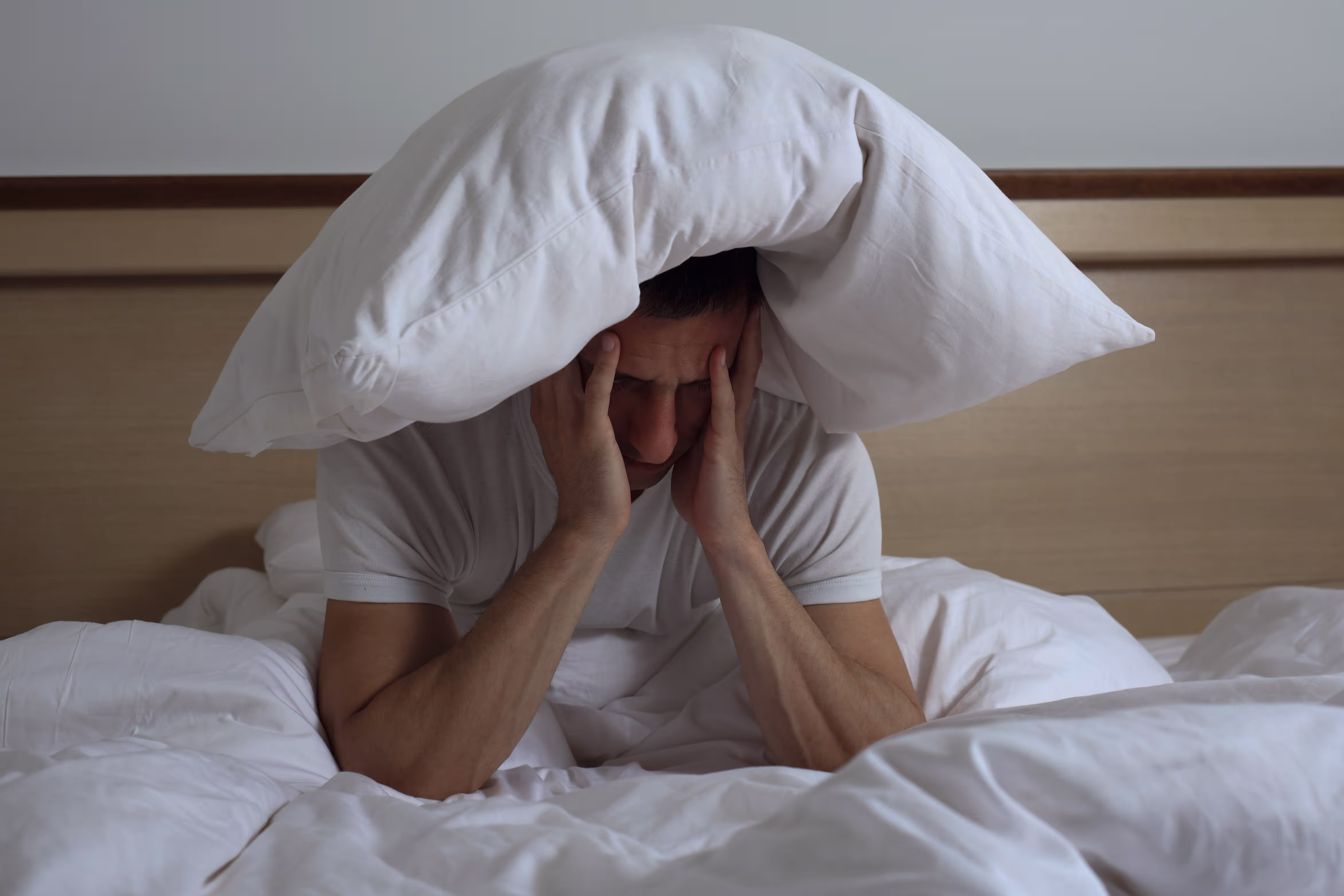 Homem sentado na cama, cansado enquanto segura o rosto com as mãos e uma almofada pousada acima da cabeça.