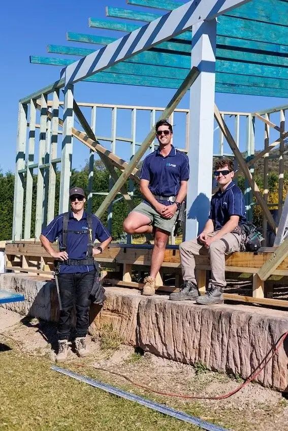 Three construction workers wearing navy shirts and sunglasses posing at a wooden building frame under a clear blue sky.
