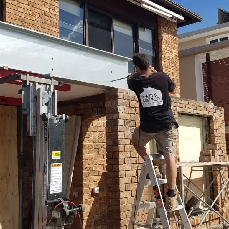 Construction worker in a black shirt and khaki shorts drilling into a white beam on a brick building while standing on a ladder.