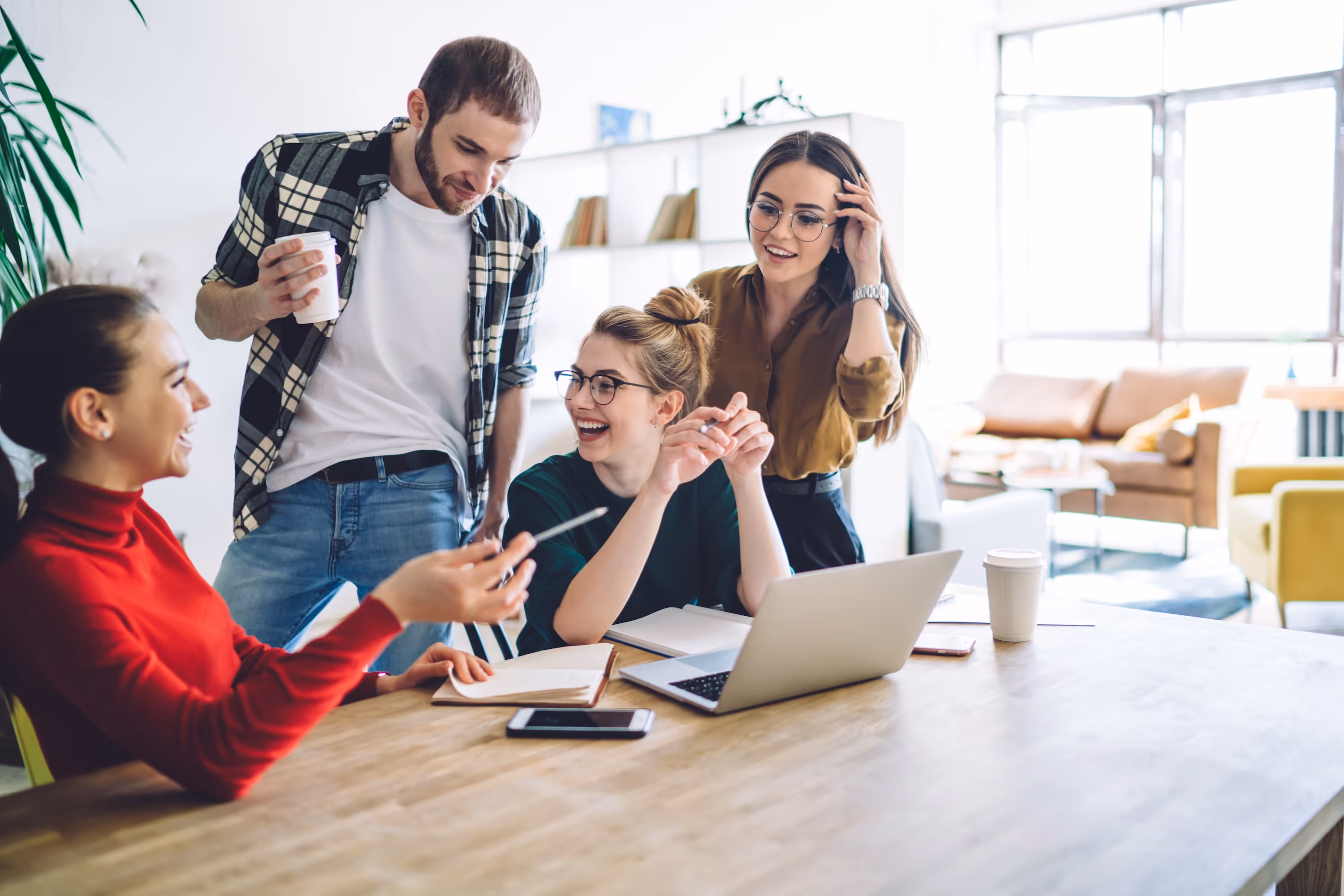 Four smiling students working together and chatting around a laptop at a communal table, representing the social and study-friendly spaces at ArtHaus Telegraph in Berkeley, CA.
