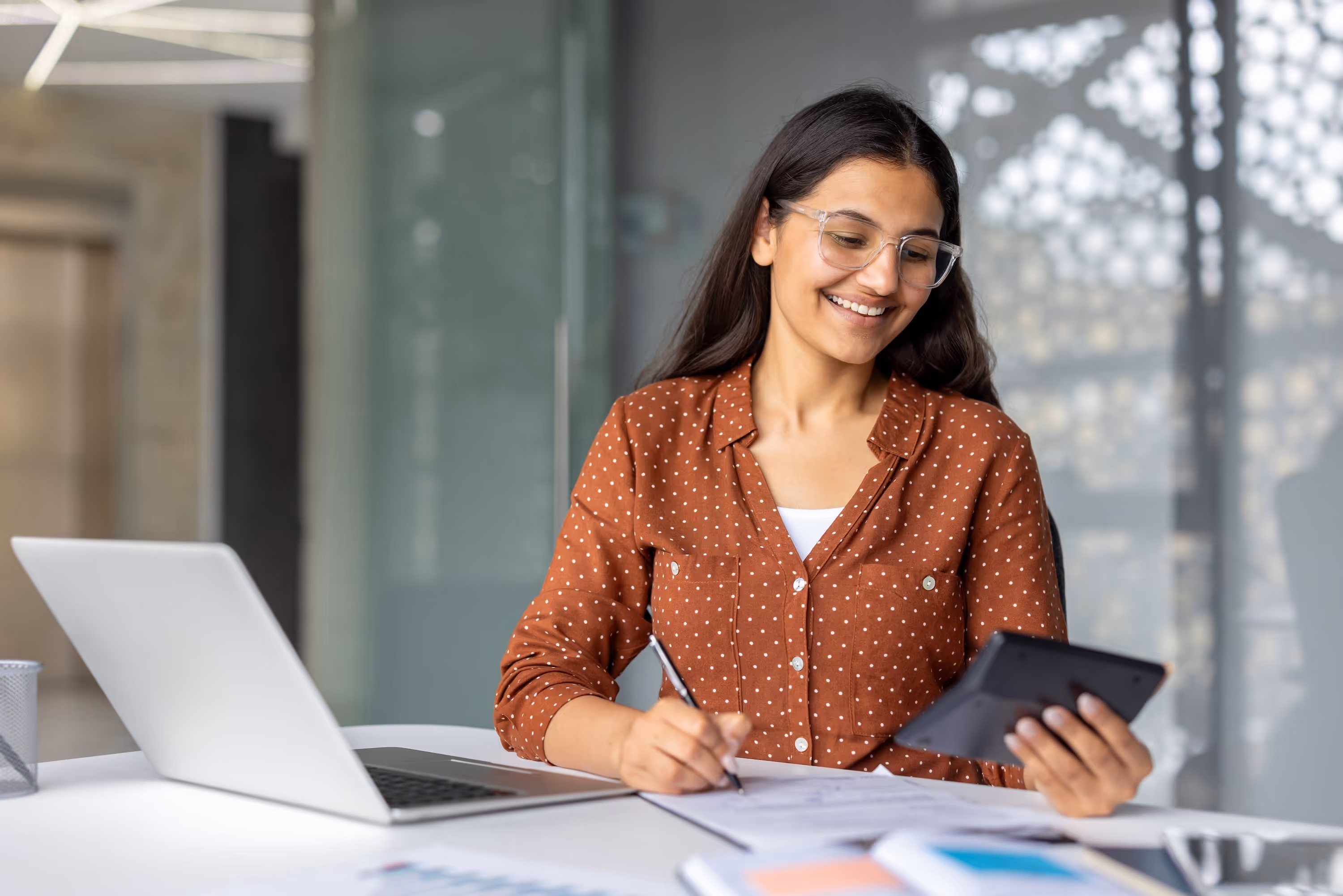 Professional woman in a office smiling while writing notes and using a calculator.