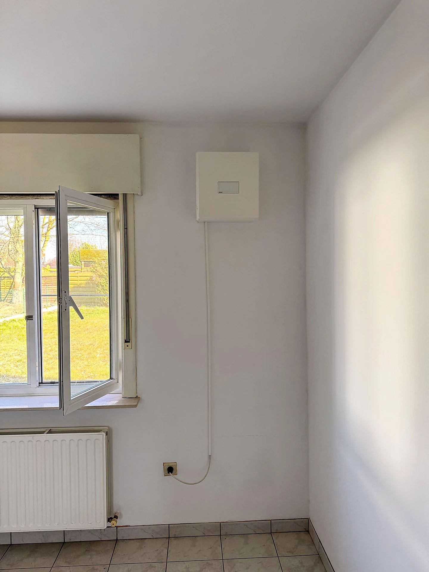 De ventilatie-unit naast het open raam met tuinzicht
