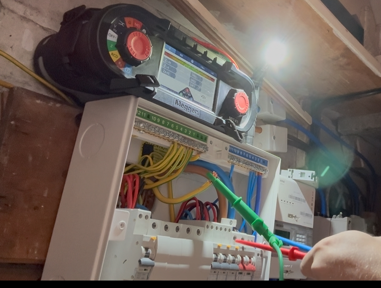 Electrical testing device connected to a consumer unit with multiple colored wires inside an industrial or utility room.