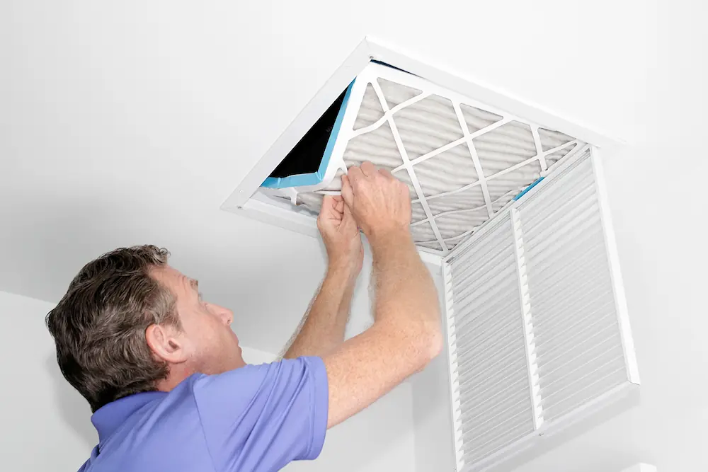 Man in a blue shirt replacing or inspecting an air filter in a ceiling vent.