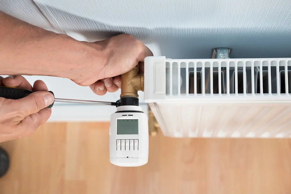 Hands using a screwdriver to install or adjust a thermostat valve on a white radiator attached to a wall.