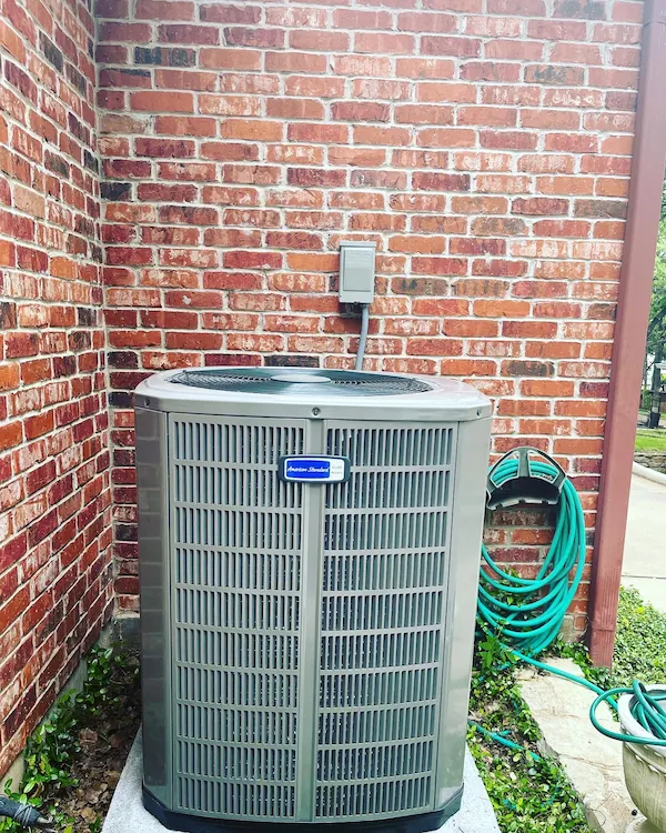 Outdoor air conditioning unit installed on a concrete slab against red brick walls with a green garden hose coiled on a wall-mounted holder nearby.