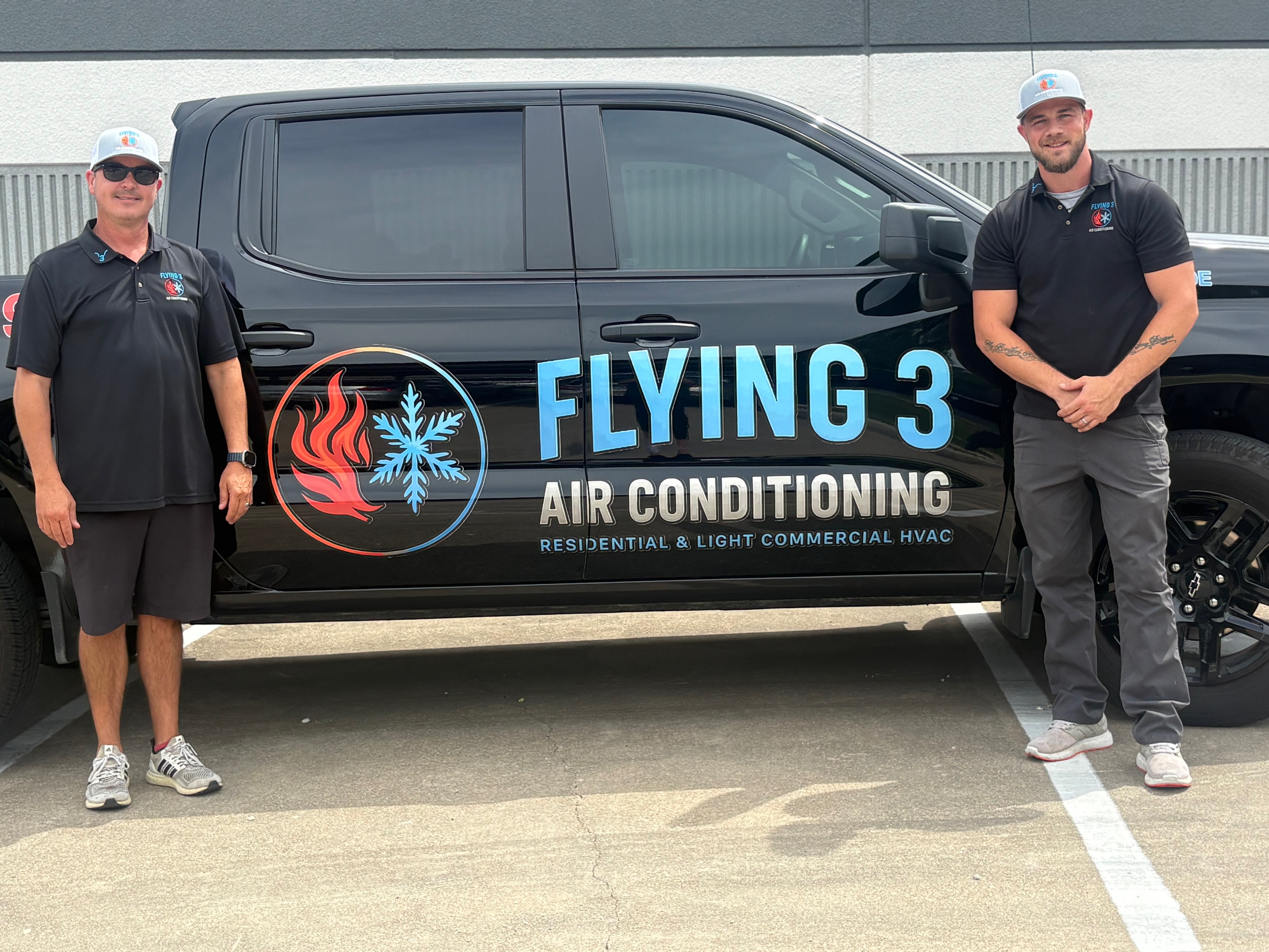 Two men in black shirts and caps stand beside a black truck with Flying 3 Air Conditioning logo.