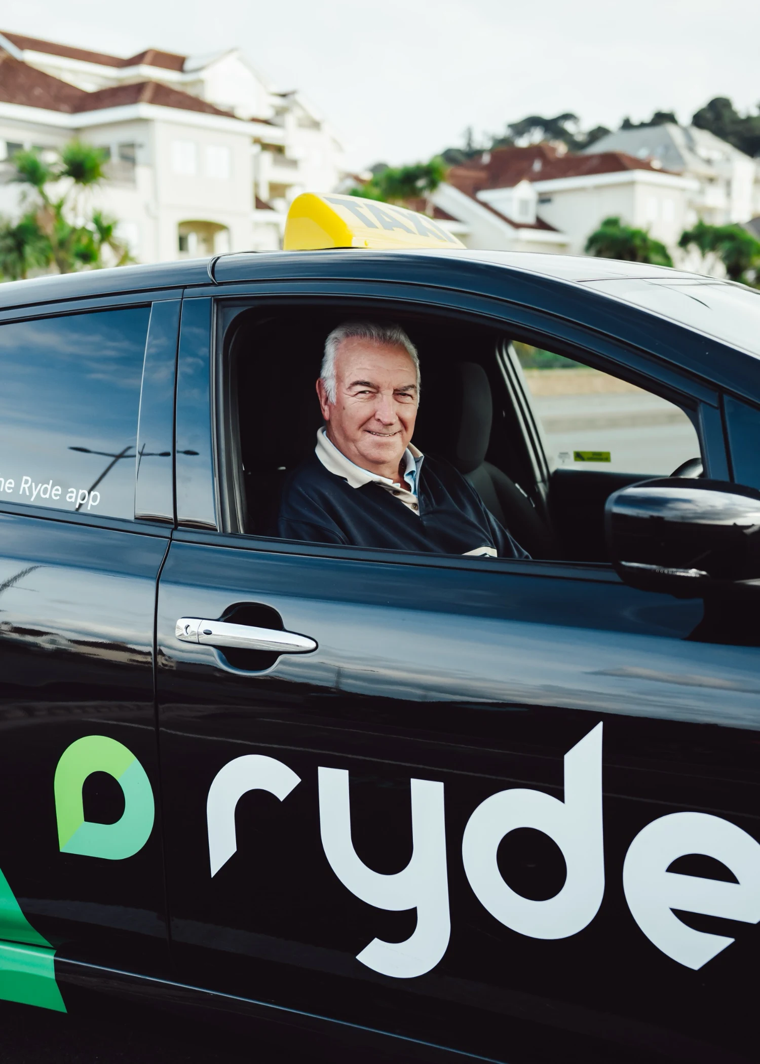 Smiling driver in black Ryde taxi with residential area in background