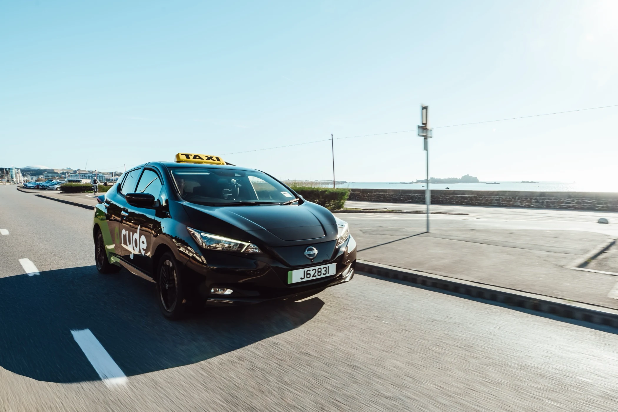 Black Nissan Leaf taxi driving on coastal road with yellow taxi sign