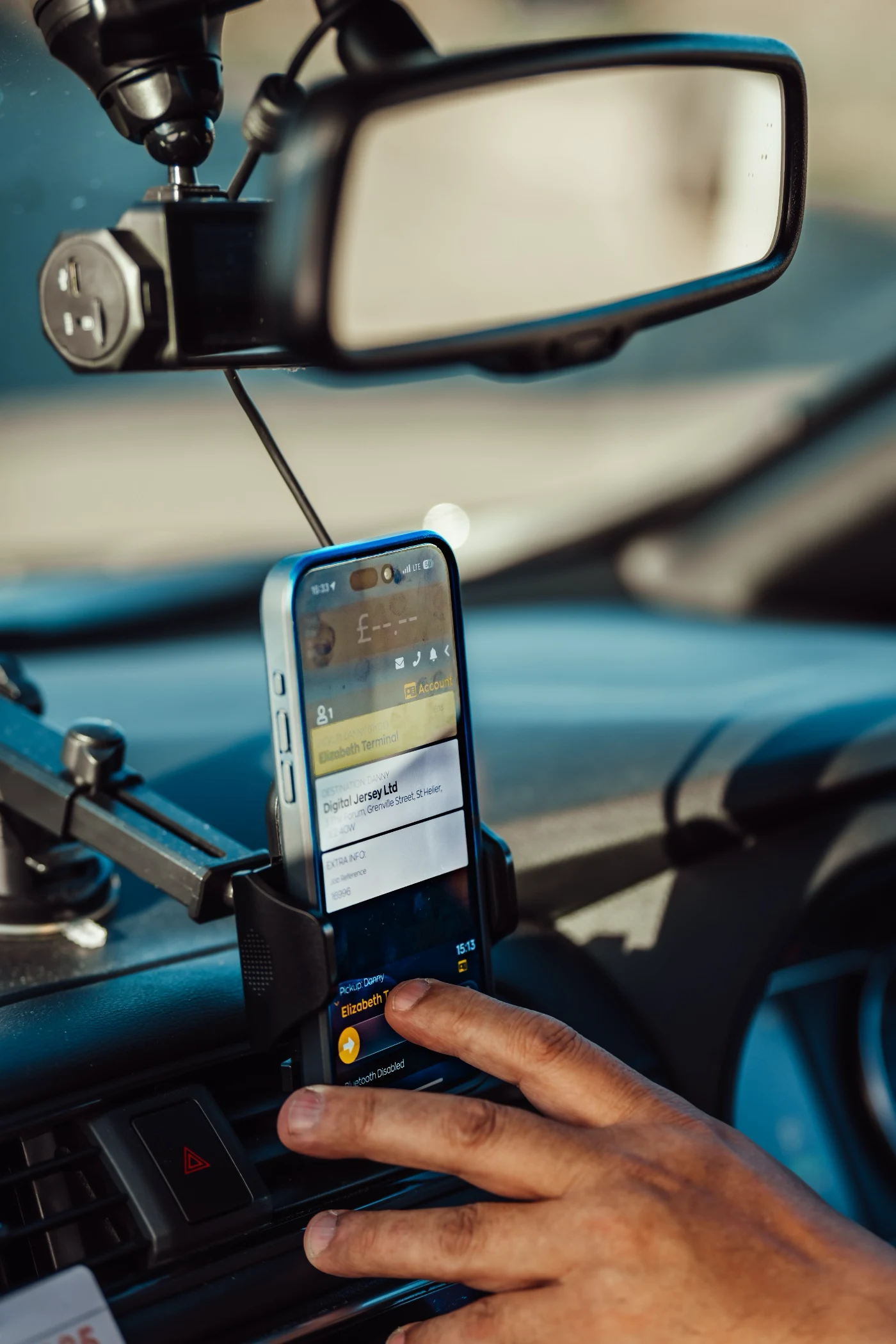 Person's hand interacting with a smartphone mounted on a car dashboard, displaying a delivery or transport app with pickup and destination details.