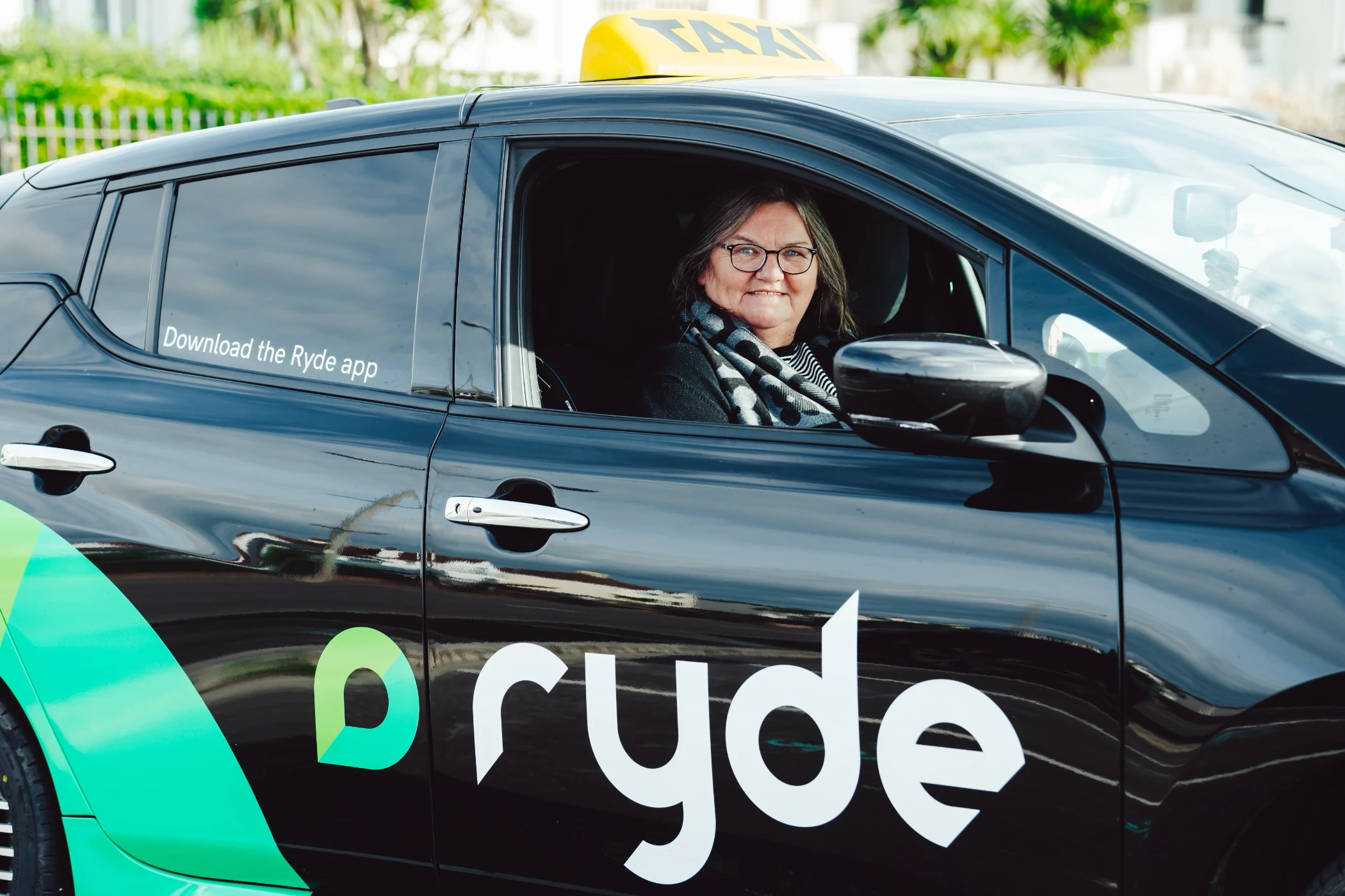 Smiling woman with glasses sitting in the driver's seat of a black Ryde taxi with a yellow taxi sign on the roof.