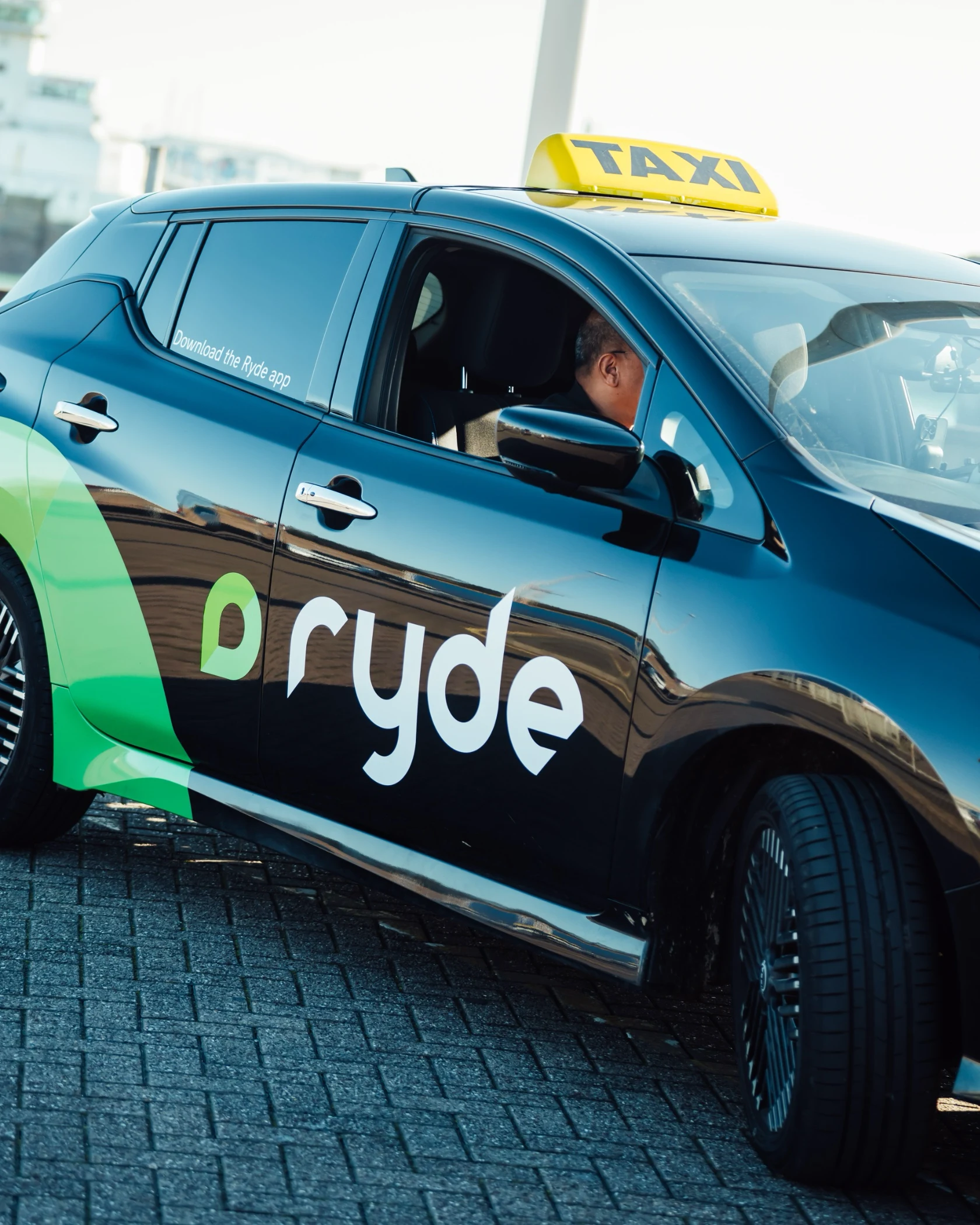 Black Ryde taxi car with green logo and a driver visible inside, parked on a paved surface.