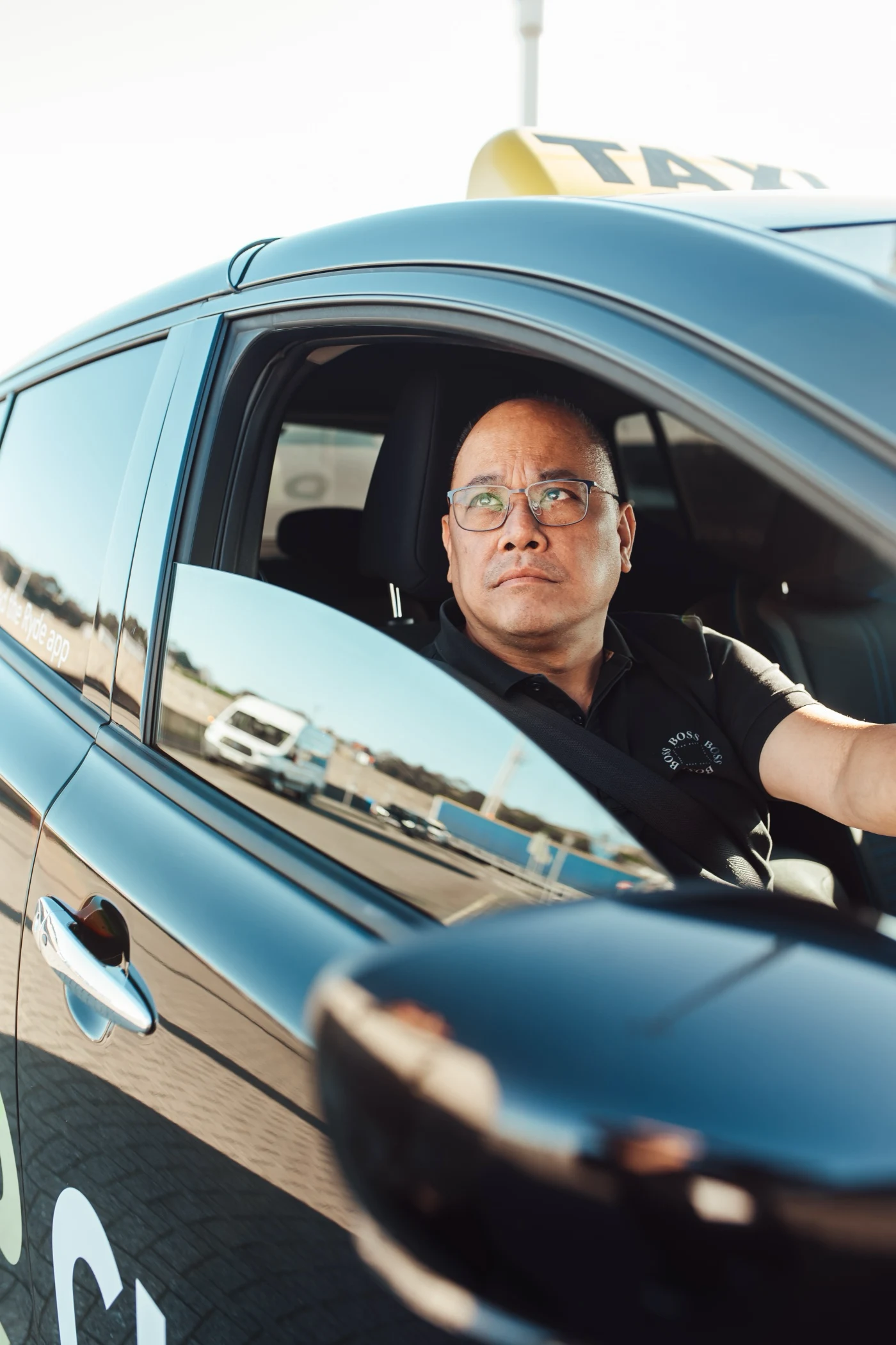 Taxi driver wearing glasses and a black shirt, looking attentively out of the car window.
