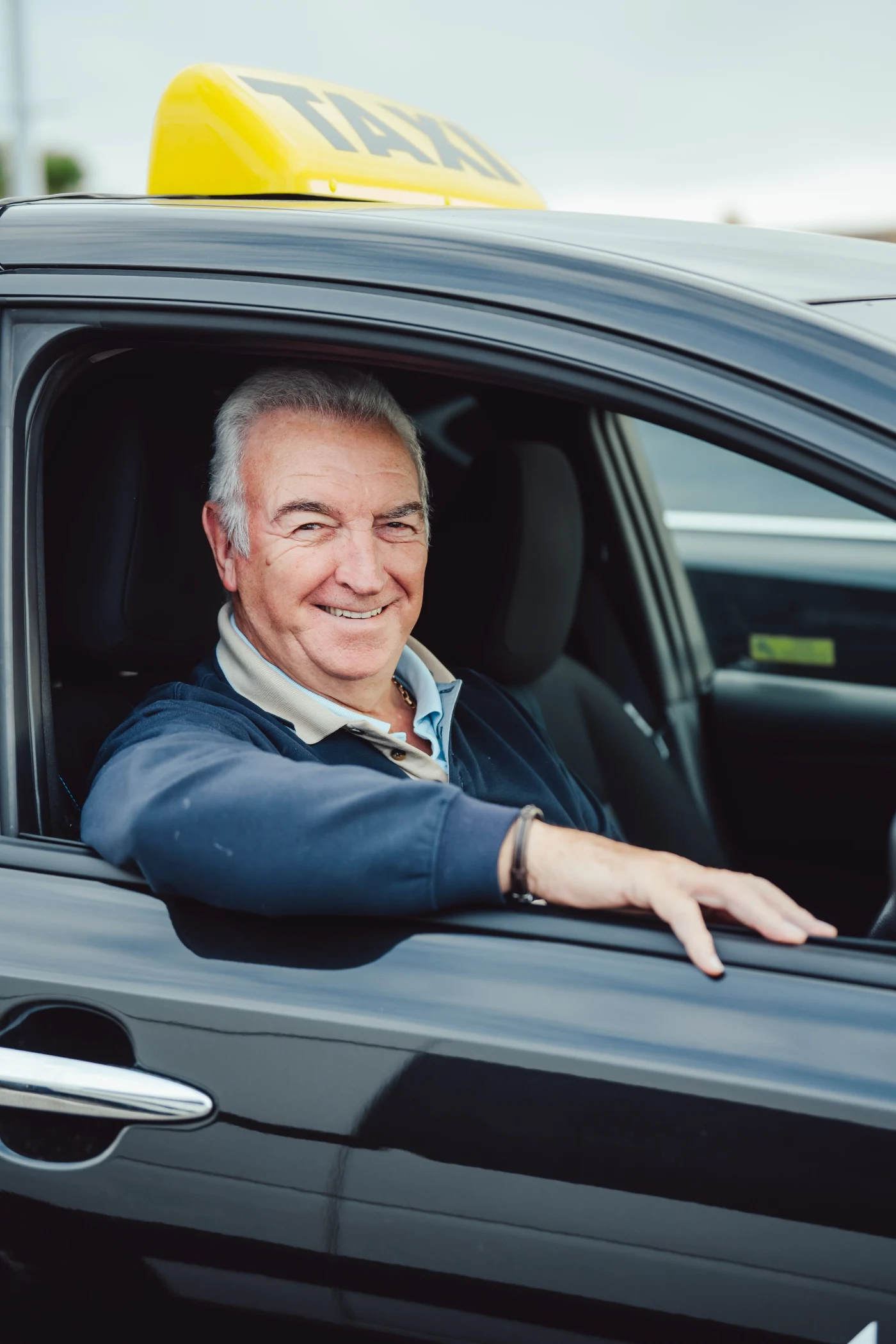 Smiling elderly man sitting in the driver seat of a black taxi with his arm resting on the open window.