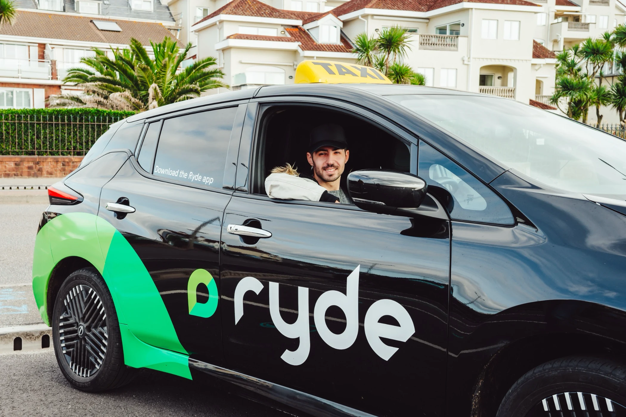 Man smiling inside a black Ryde taxi with green branding and a yellow taxi sign on top, parked on a street with residential buildings in the background.