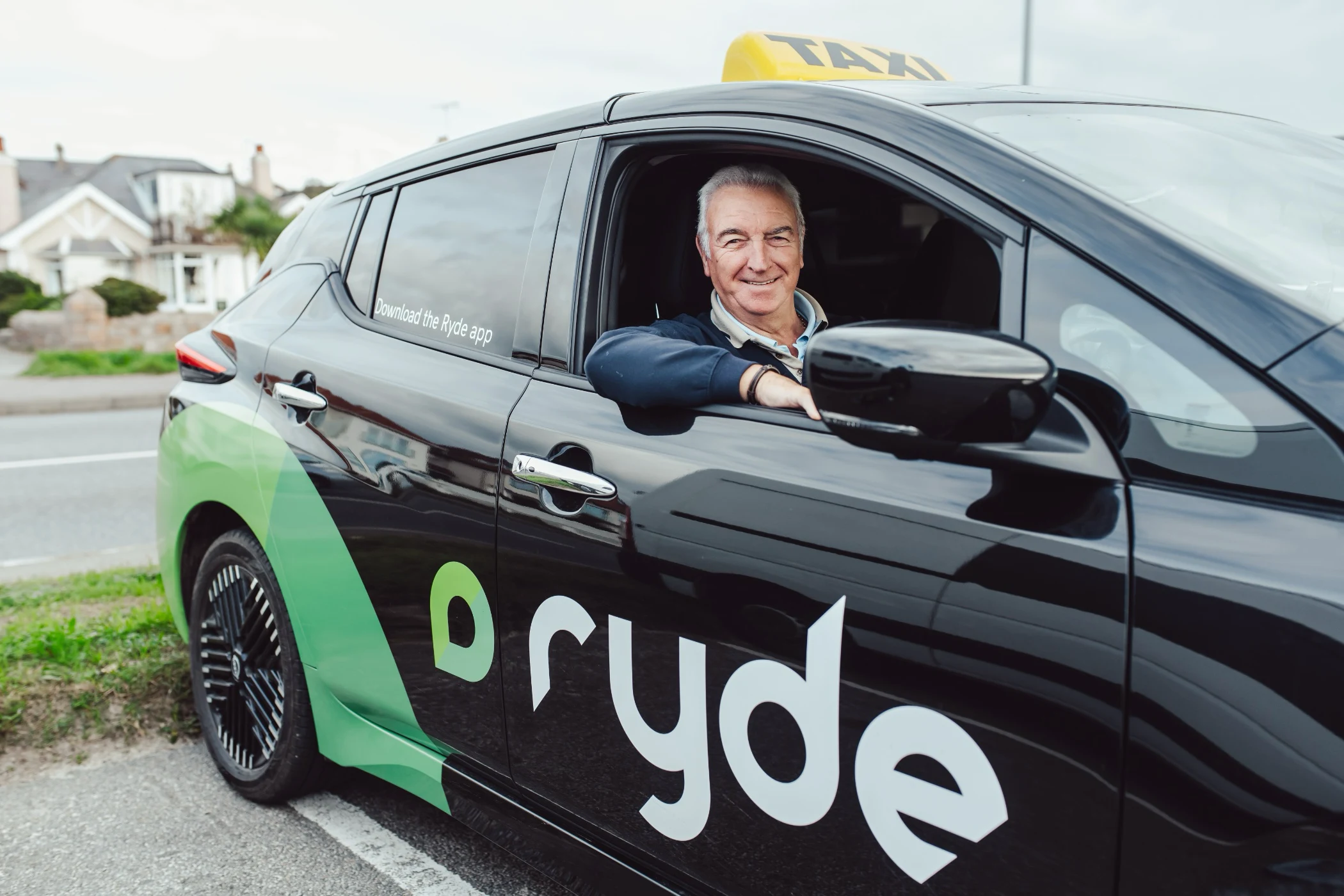 Smiling middle-aged man sitting in the driver seat of a black Ryde taxi car with green logo.