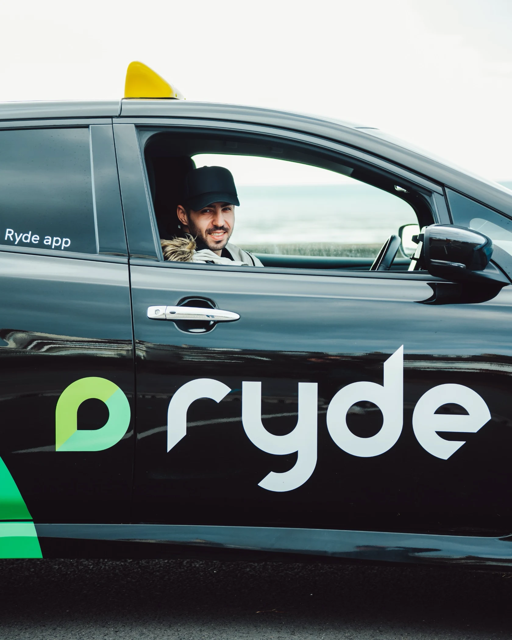 Man wearing a black cap sitting in the driver's seat of a black Ryde app taxi with a yellow roof sign.