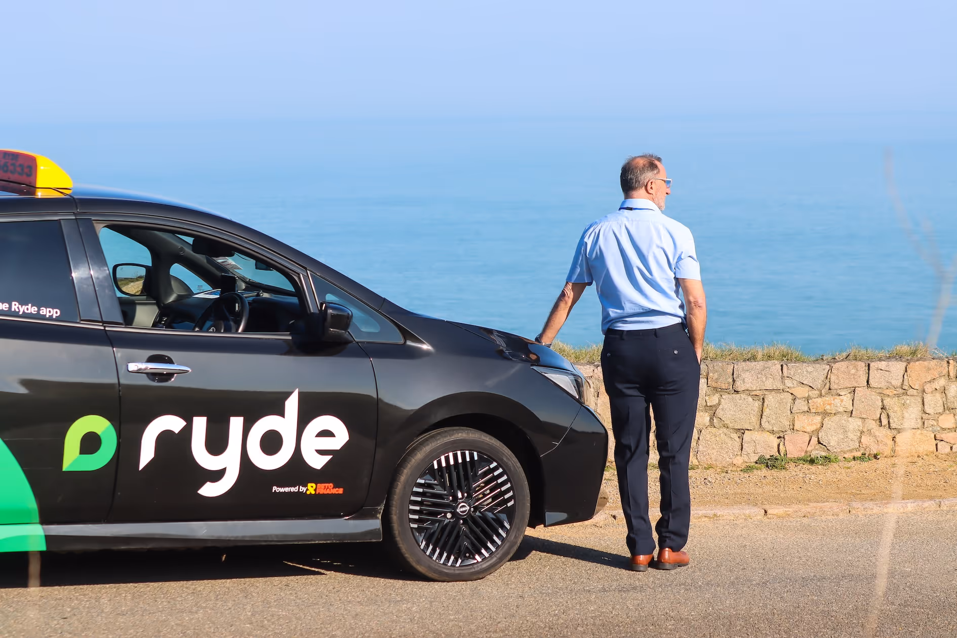 Man in blue shirt and dark pants leans on black Ryde taxi by a stone wall with the ocean in the background.