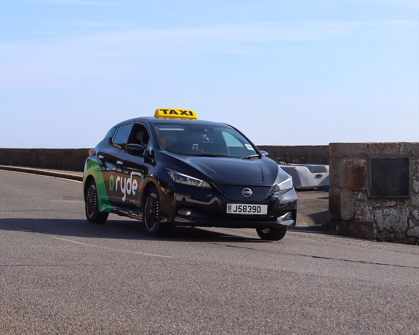 Black Nissan taxi with green Ryde logo driving on paved road near stone wall and clear sky.