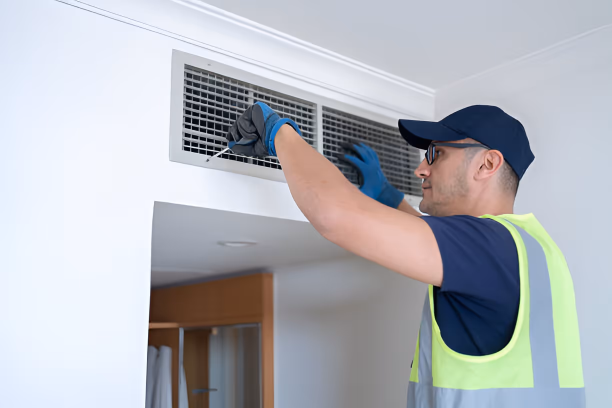 Technician wearing gloves, glasses, and a cap inspecting or repairing a wall air vent inside a room.
