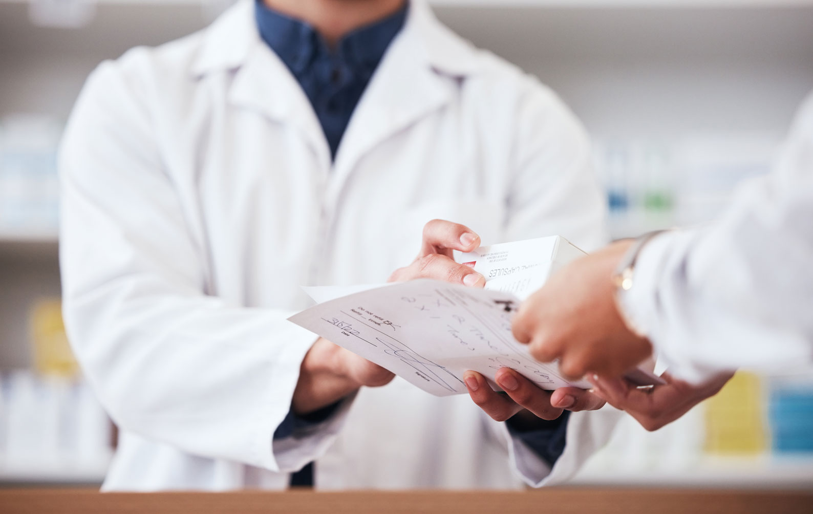 Two pharmacists exchanging a prescription paper and a medication box at a pharmacy counter.