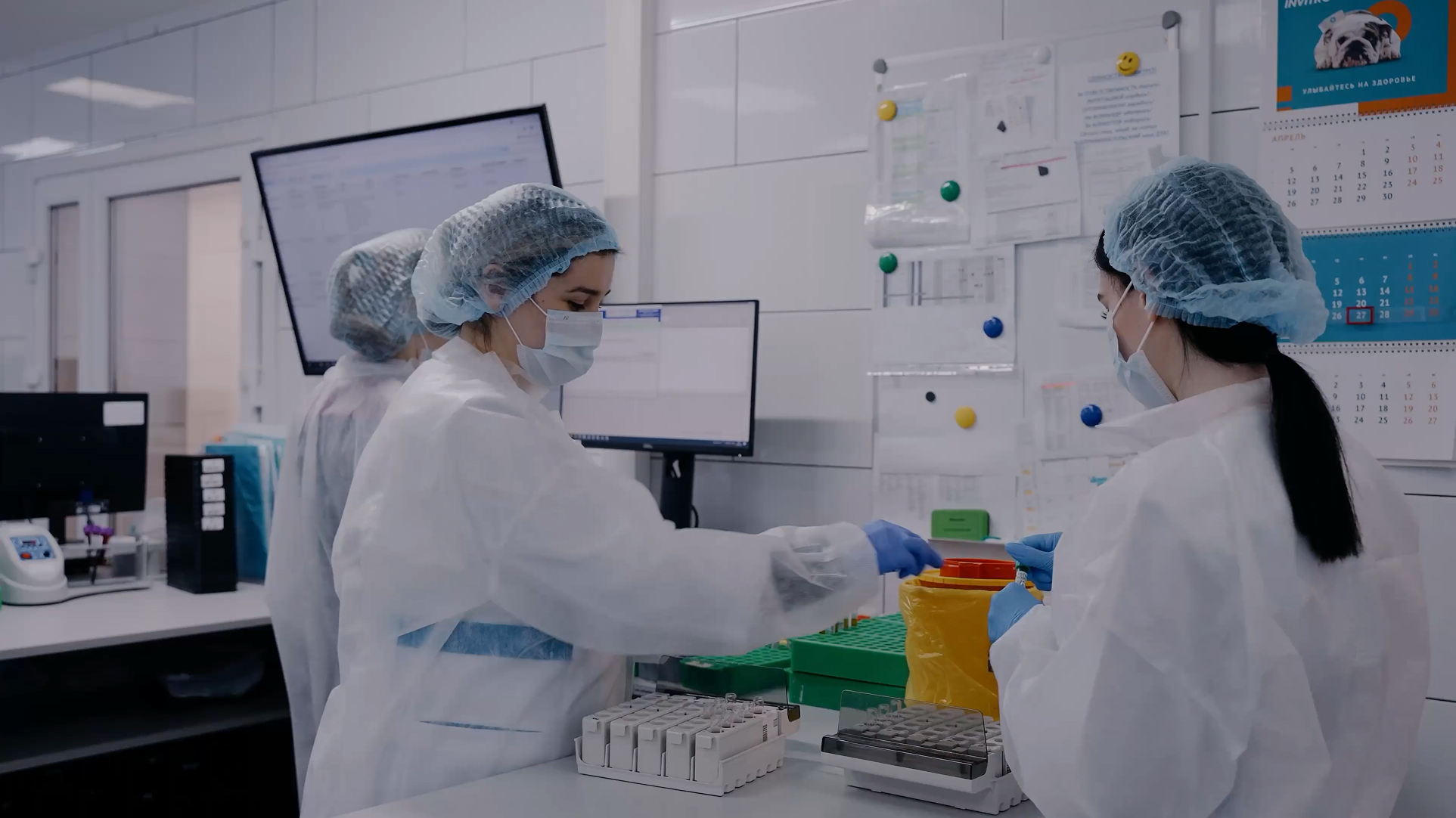 Two laboratory workers wearing protective gear in a clinical lab handling test tubes near computer monitors.