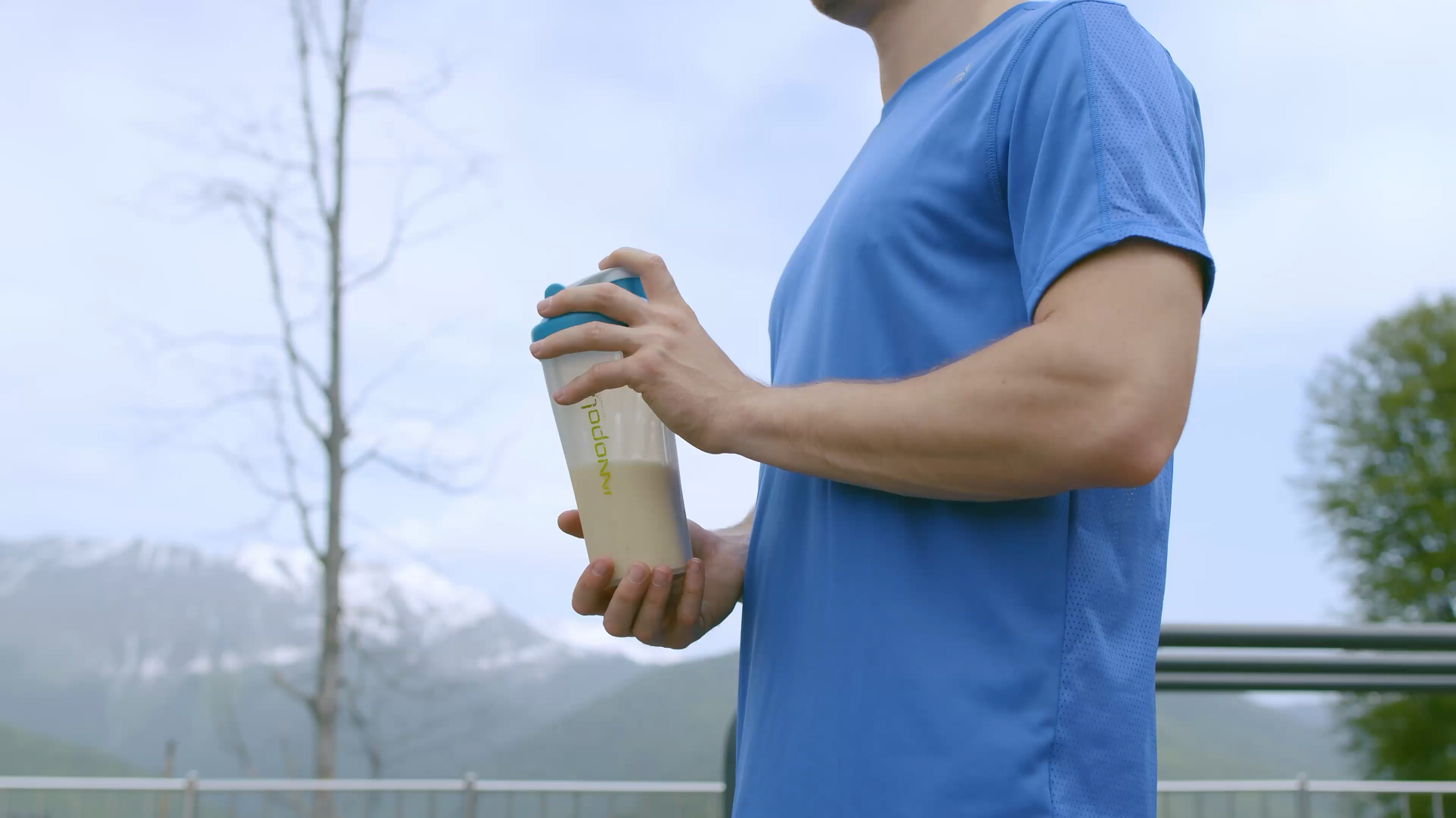 Person in blue athletic shirt holding a clear shaker bottle with beige liquid outdoors with mountains in the background.