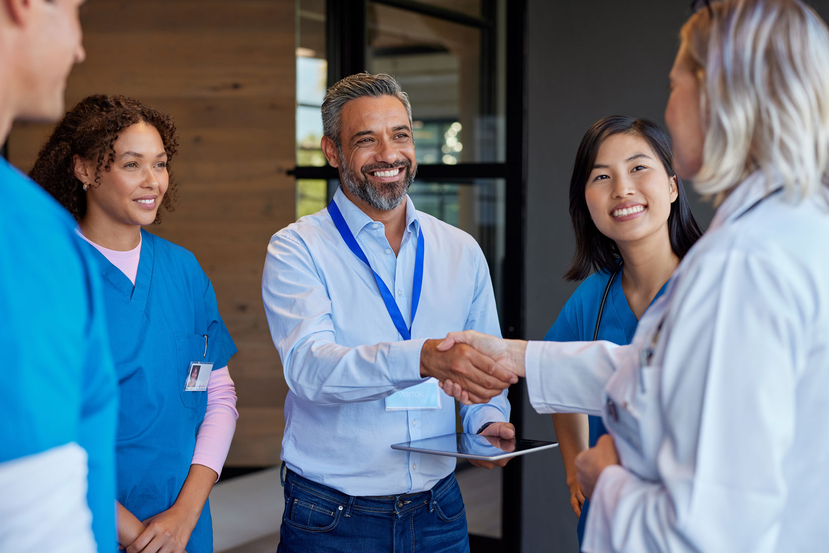 Man with visitor badge shaking hands with a female doctor while two nurses look on smiling.