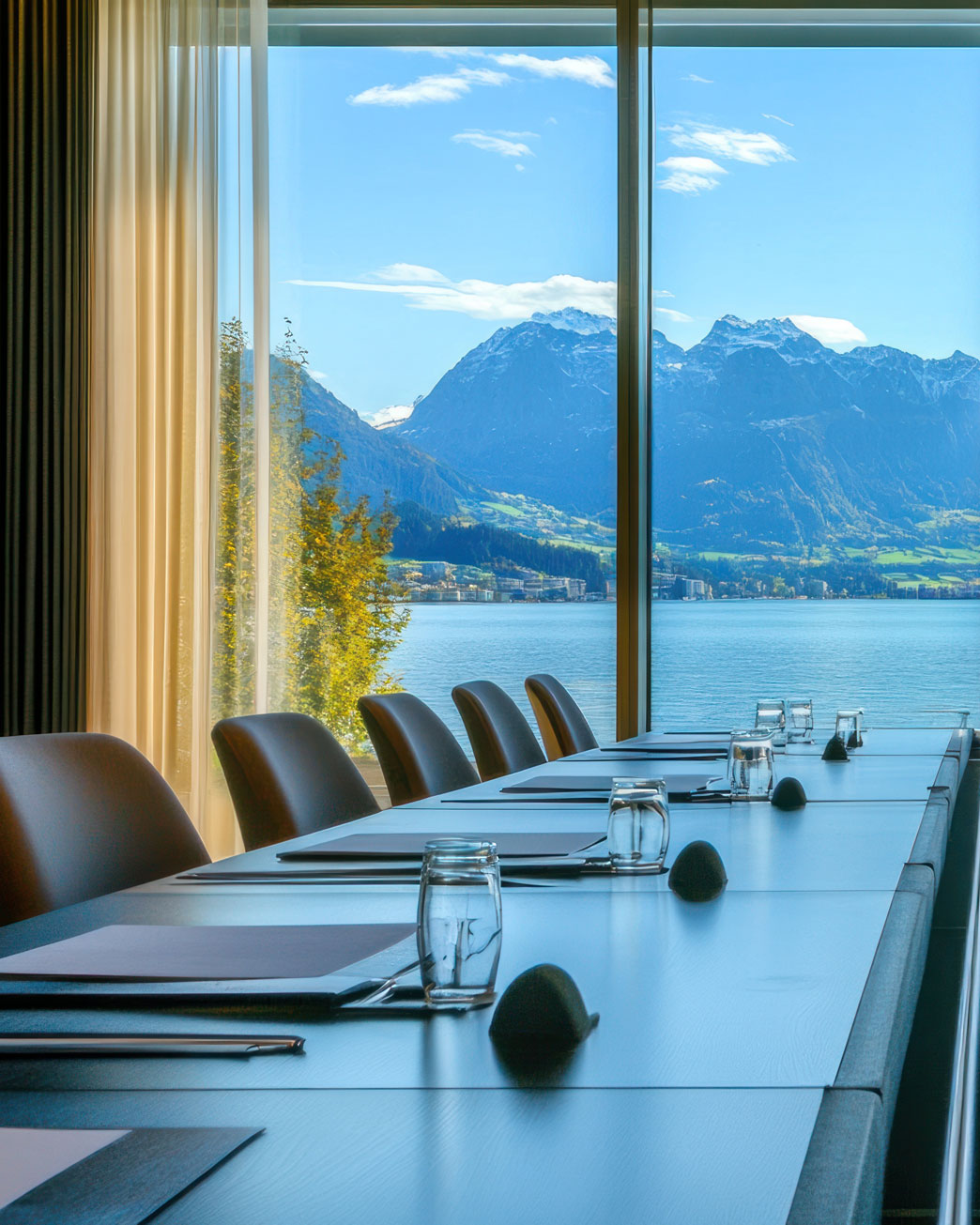 Modern conference room table set with glasses, notebooks, and pens, overlooking a lake and mountain view through large windows.