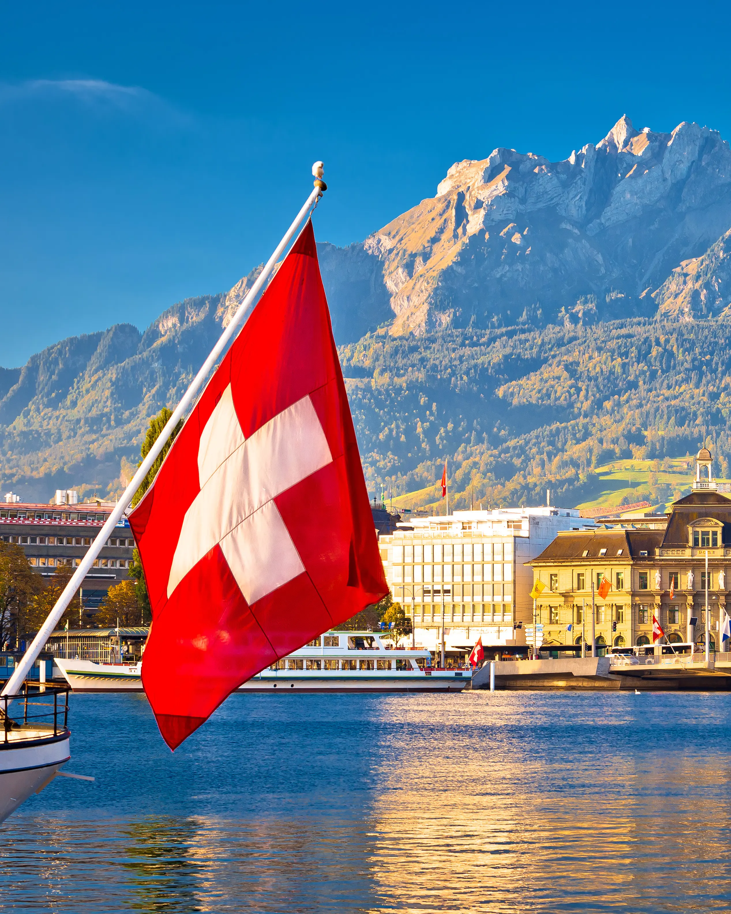 Swiss flag waving on a boat with a lakeside town and mountains in the background under a clear blue sky.