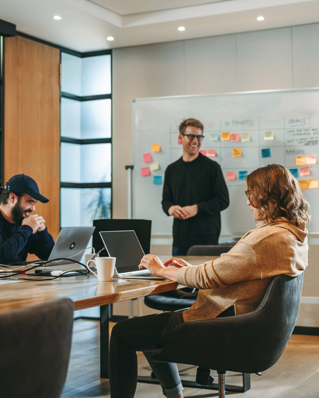 Three young professionals collaborating in a modern office with laptops and a whiteboard covered in colorful sticky notes.