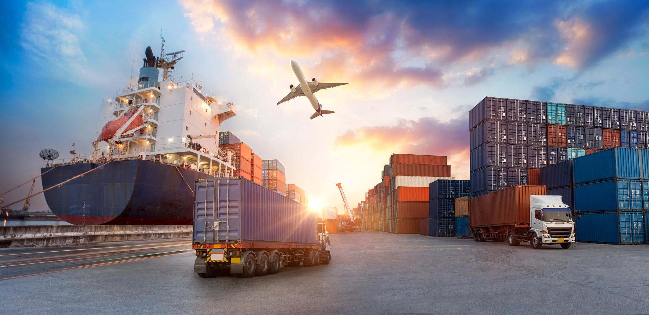 Cargo trucks and container ship at a shipping port with an airplane flying overhead during sunset.