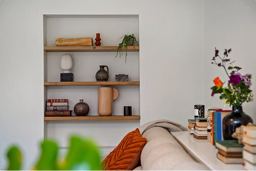 Cattenburg interieur design. Ingebouwde muurplanken met decoratieve potten, boeken en planten naast een beige bank met oranje kussen en boeken op de leuning.