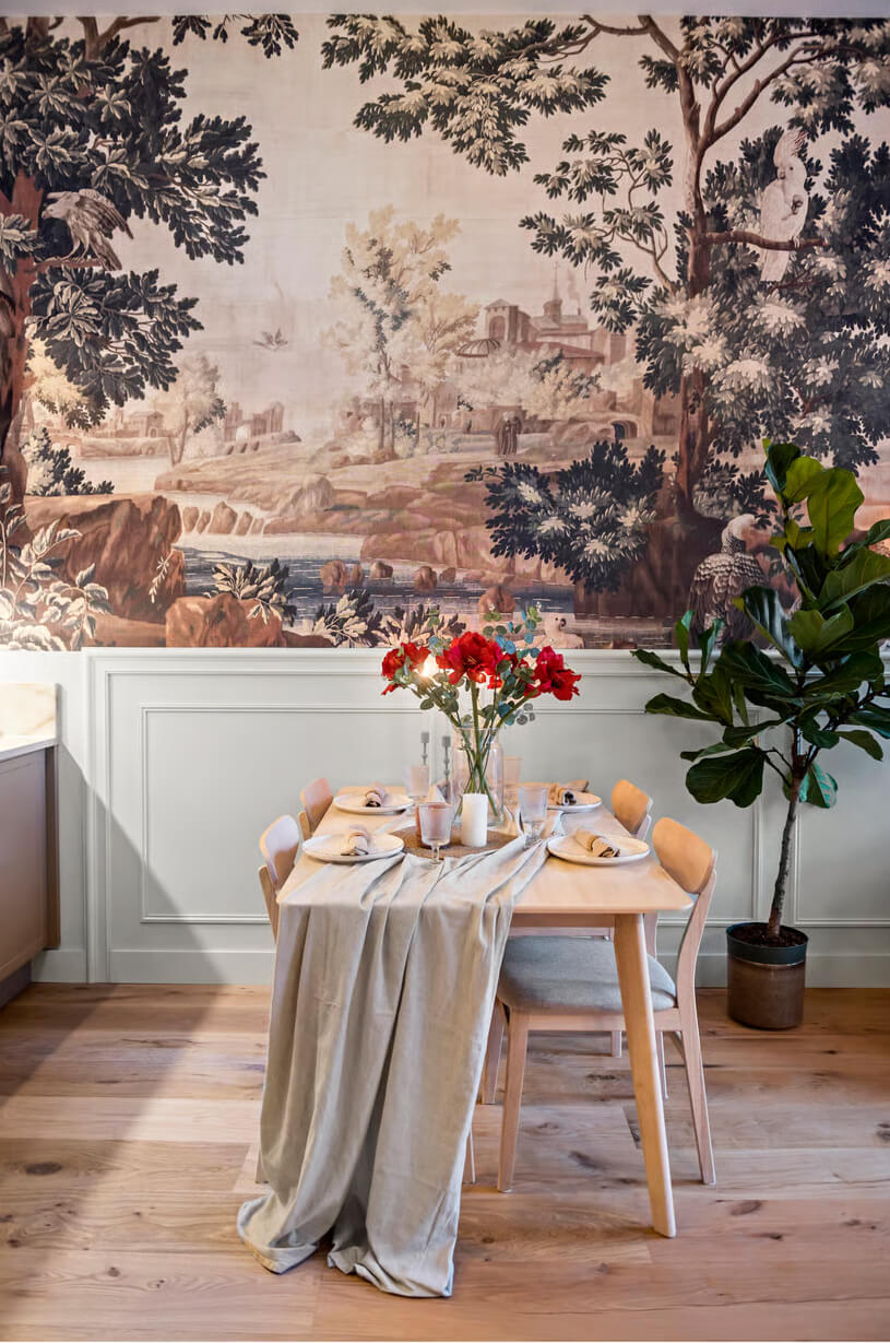 Cattenburg interieur design. Gezette eettafel met beige tafelloper, rode bloemen in vaas en servies, voor een decoratieve muurschildering met bos- en waterelementen.