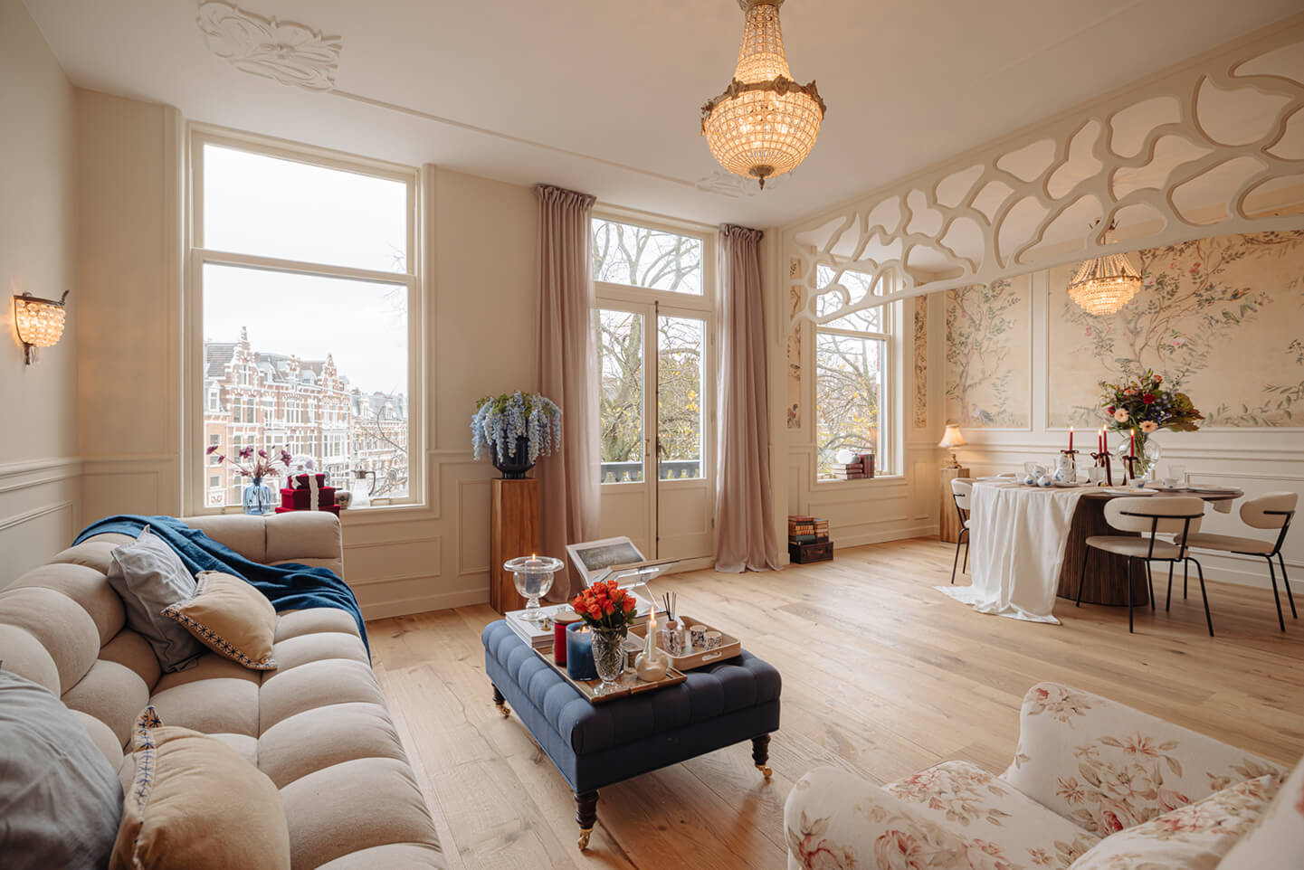 Lichte woonkamer met beige bank, blauwe poef met decoraties, bloemen en een eettafel met kaarsen onder een kroonluchter.