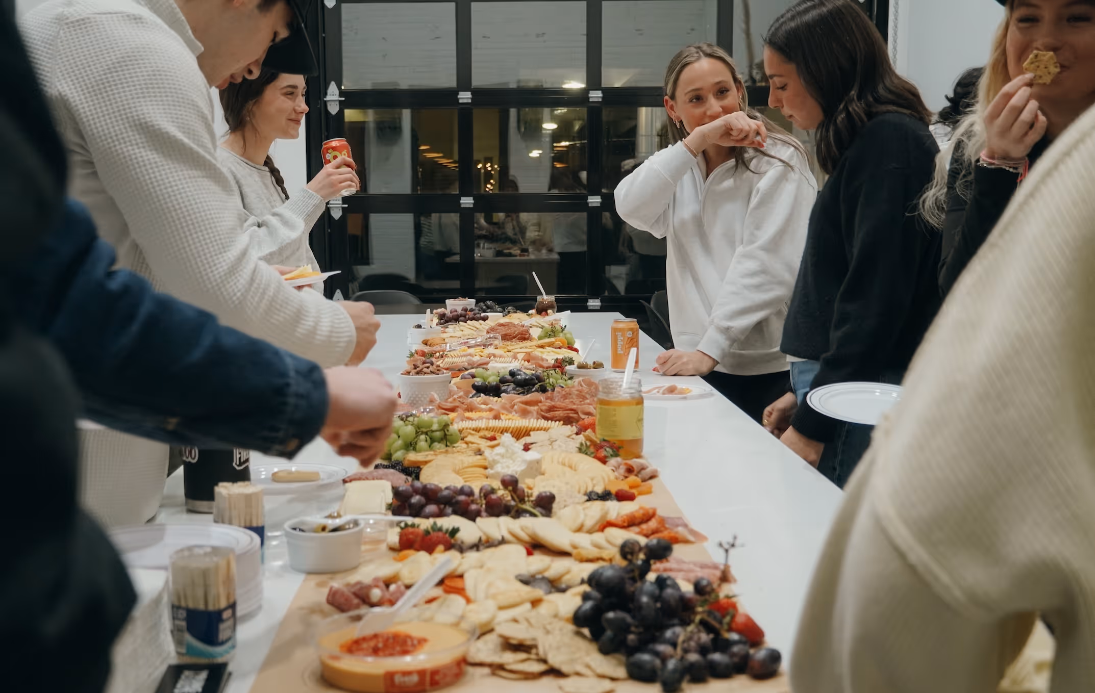 Group of young adults gathered around a long table with assorted charcuterie, cheese, fruits, and snacks.